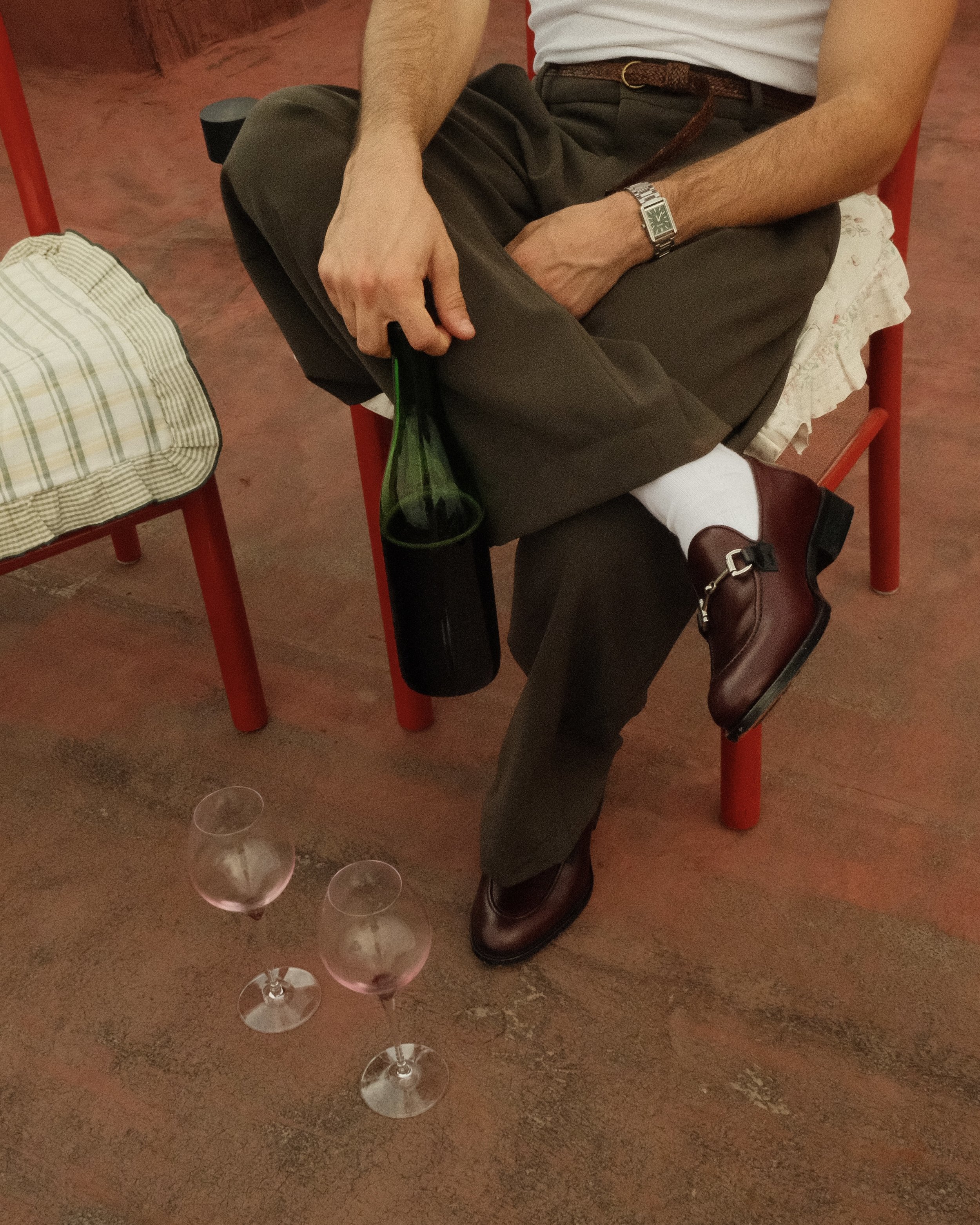 Person auf einem roten Stuhl sitzend, mit einer grünen Flasche; drei Weingläser auf dem Boden; trägt braune Schuhe und eine Armbanduhr.