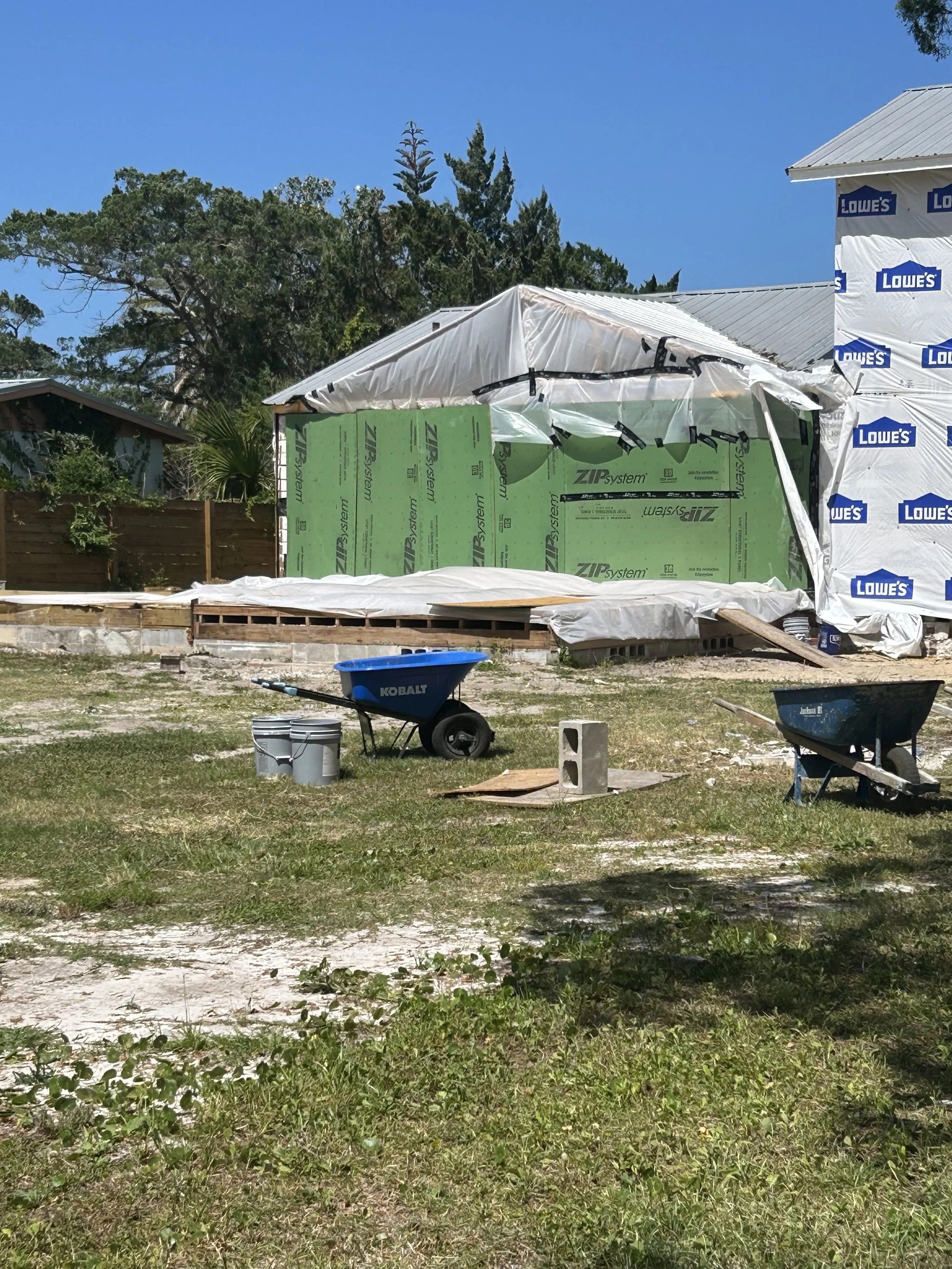 A house under construction with partially completed walls covered with green and white building wrap, and tools such as wheelbarrows and buckets in the foreground, set on a grassy lot. Located in the City of Saint Augustine.