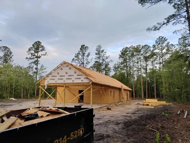 Under construction wooden house in a wooded area with construction materials and debris surrounding it. This home is located in Flagler Estates in St. John's County, FL. We are one of the local custom builders who build in that area.