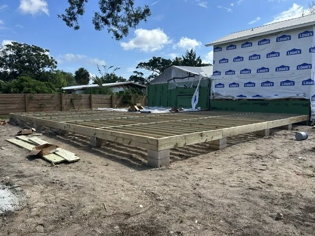 Construction site with a wooden foundation frame on dirt, surrounded by construction materials and a completed building wrapped in Lowe's house wrap in the background. This house is located in the City of Saint Augustine in the heart of St. John's.