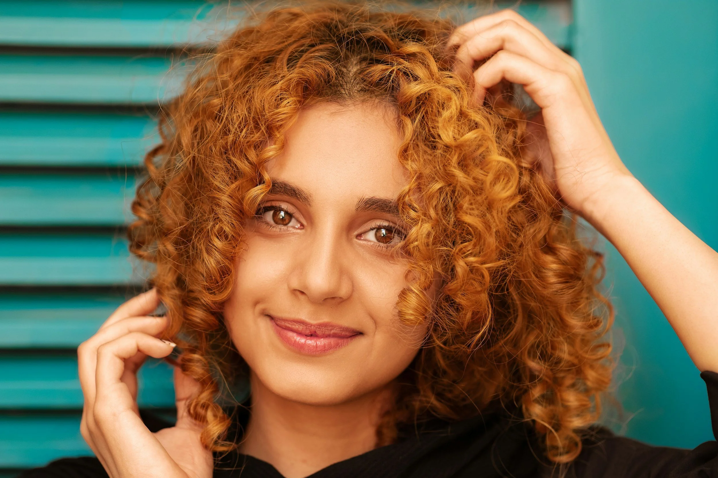 A woman with curly red hair smiling and posing with one hand near her head and the other touching her hair, against a teal background. - highlighting the clear skin of someone who visits her dermatologist to maintain healthy skin habits