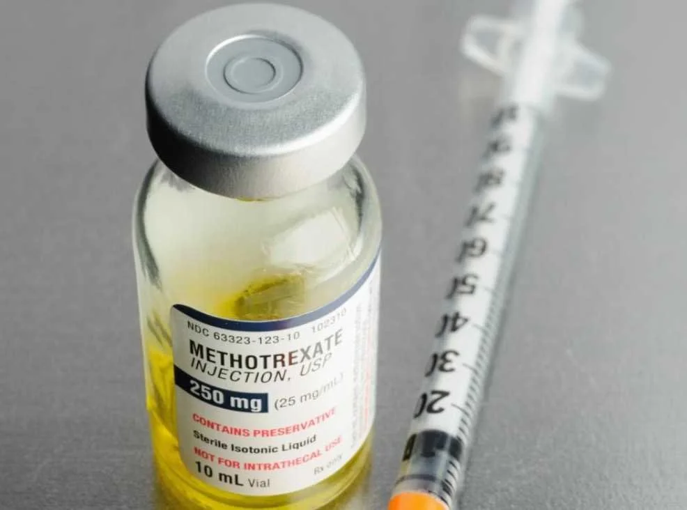 A vial of Methotrexate injection with a syringe beside it, placed on a gray surface.