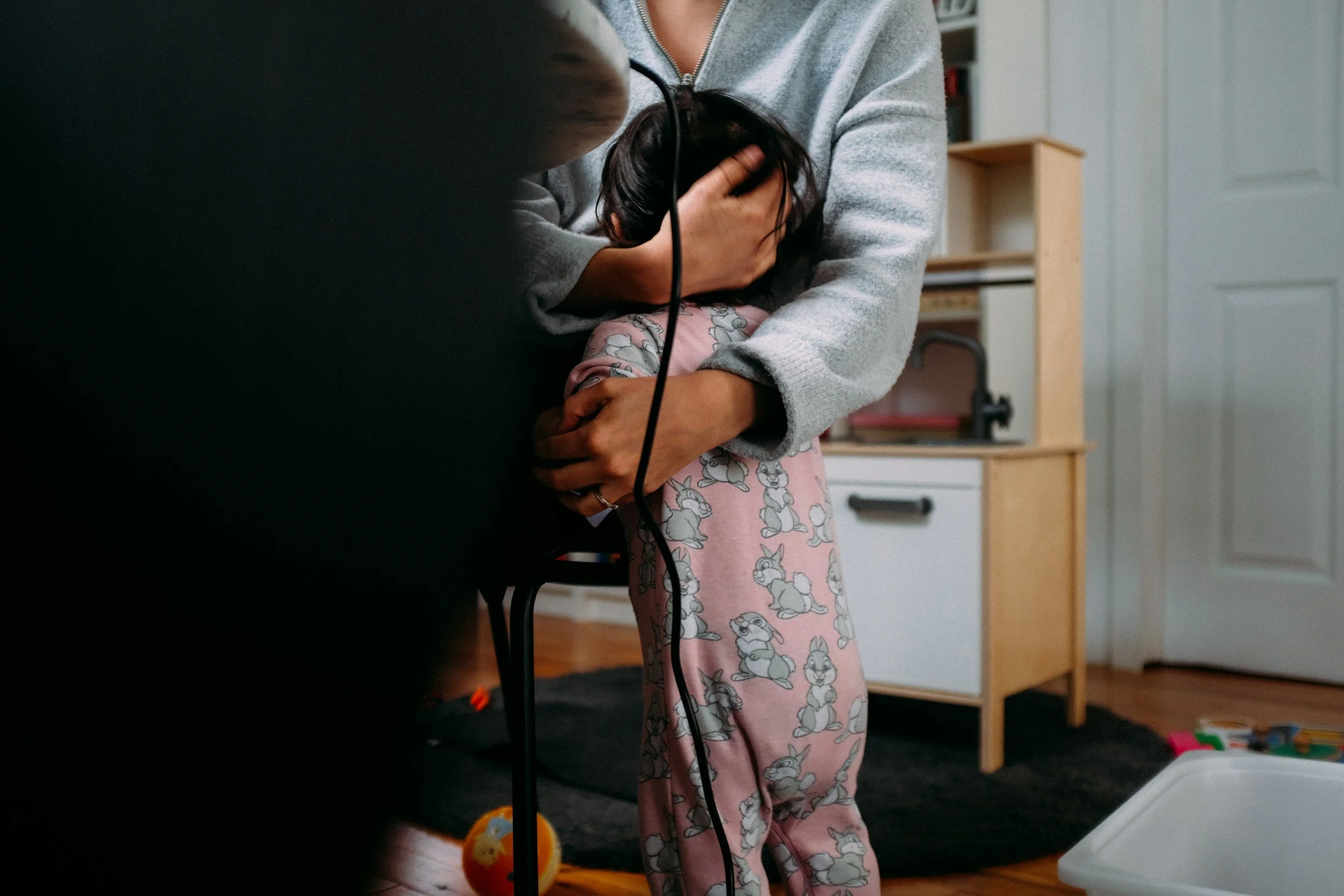 A person wearing pink pajamas with bunny prints holding a child's head while another person in a gray sweater supports the child's head with their hand. The child is facing down, and a black cord is visible. The scene appears to be in a room with a wooden floor, white door, and bookshelf in the background.