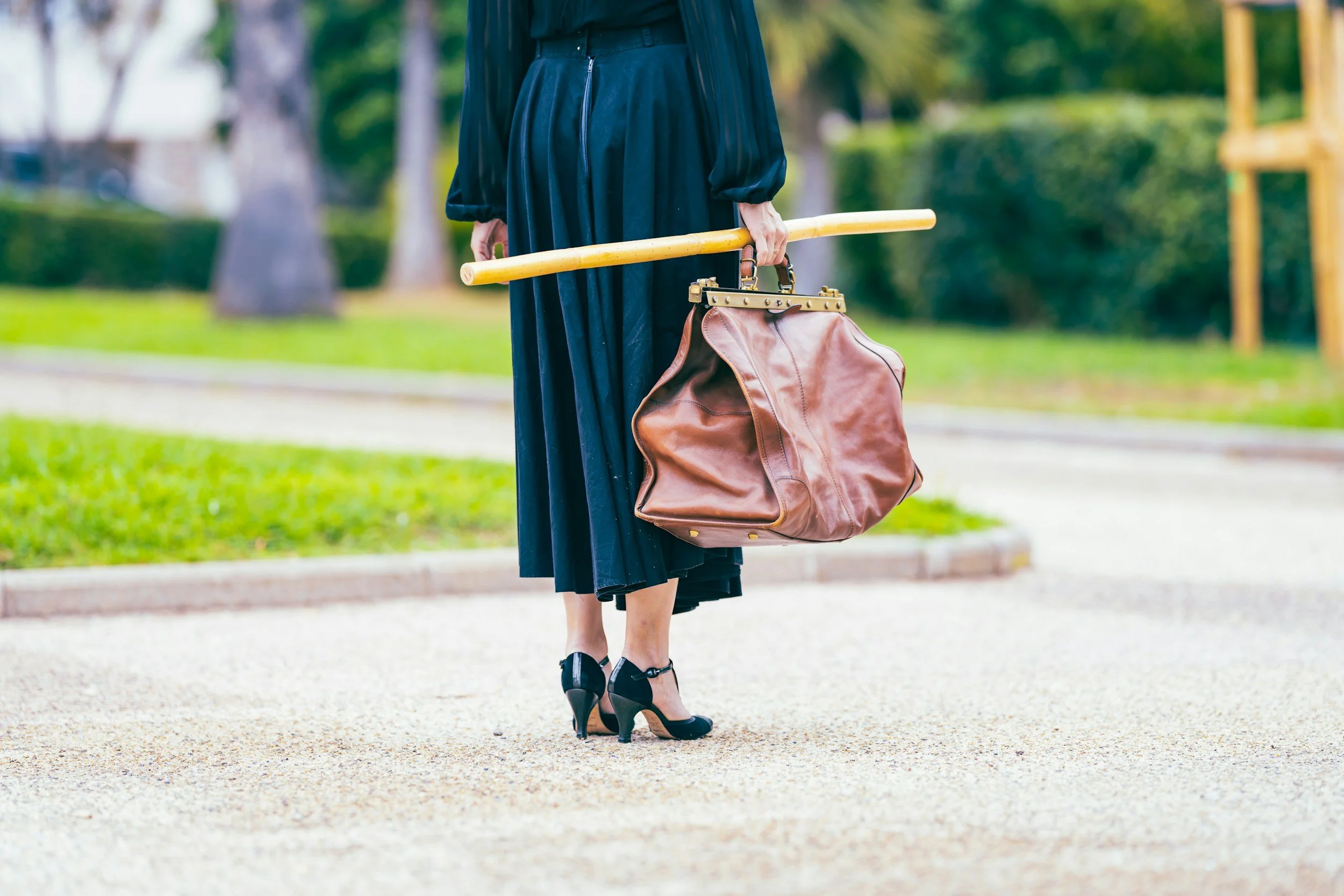 A person wearing a black dress and high heels walking outdoors in a park, holding a large brown leather bag in one hand and a yellow bamboo stick in the other.