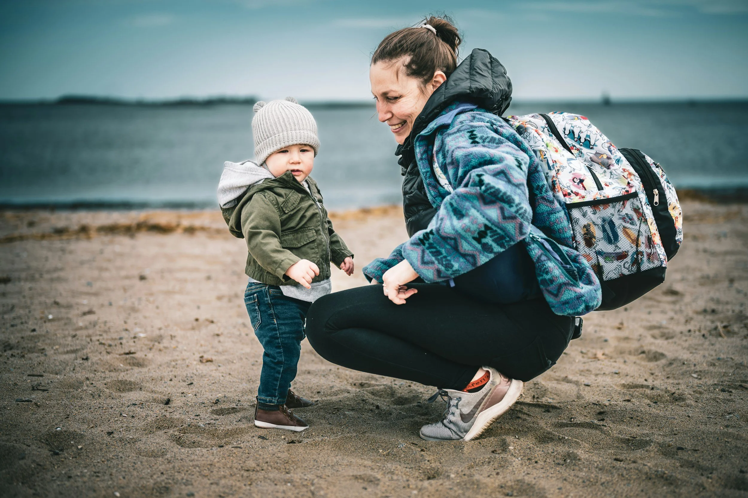 A woman crouching on a sandy beach talking to a young child. The woman has a backpack and is dressed in outdoor clothing, smiling, while the child wears a gray knit hat and a green jacket.