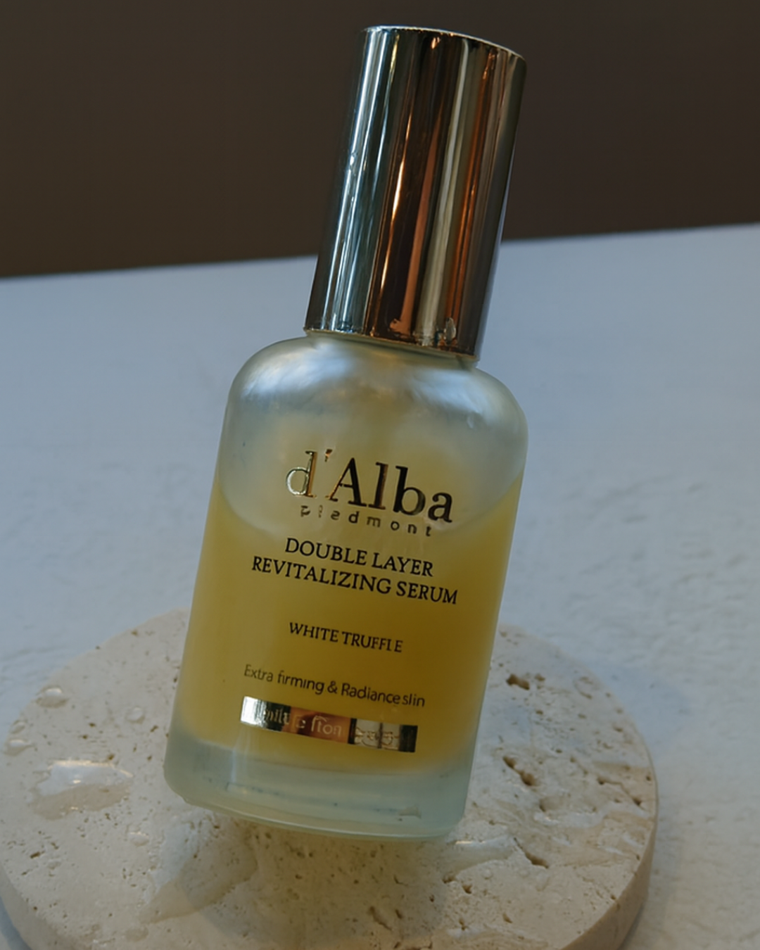 A d'Able Piedmont Double Layer Revitalizing Serum bottle with a metallic cap, filled with yellow serum, standing on a textured circular stone base against a neutral background