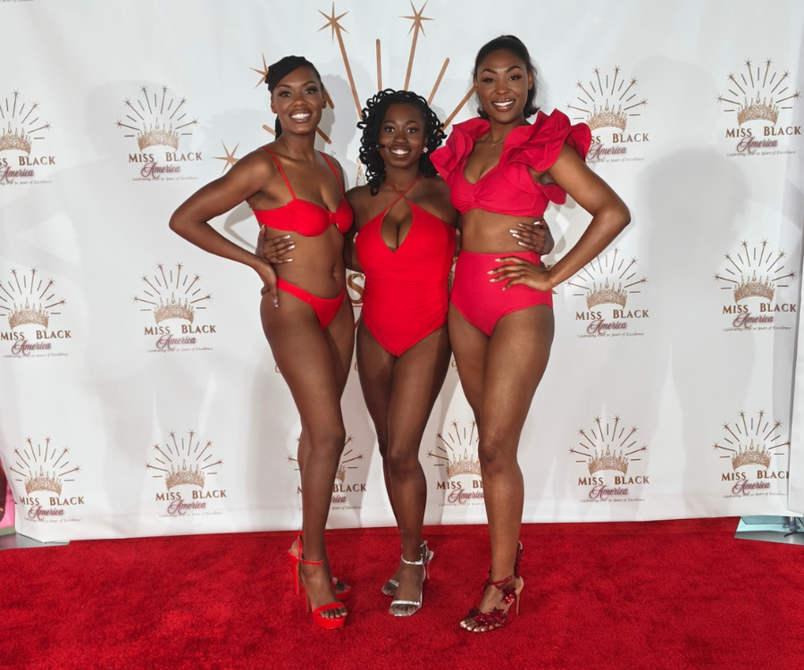 Three beautiful black women standing side by side wearing red bathing suits, standing in heels on a red carpet with a crown background