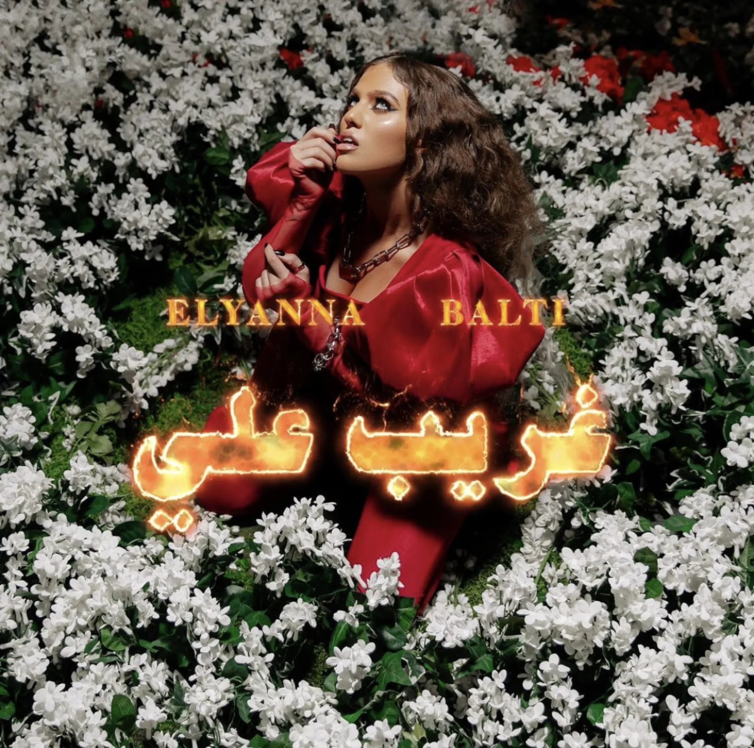 Styled by celebrity stylist and costume designer, Amanda Massi, music artist Elyanna wearing a custom red satin outfit, is sitting among white flowers with some red flowers in the background. There is glowing text in Arabic script, with the names 'El