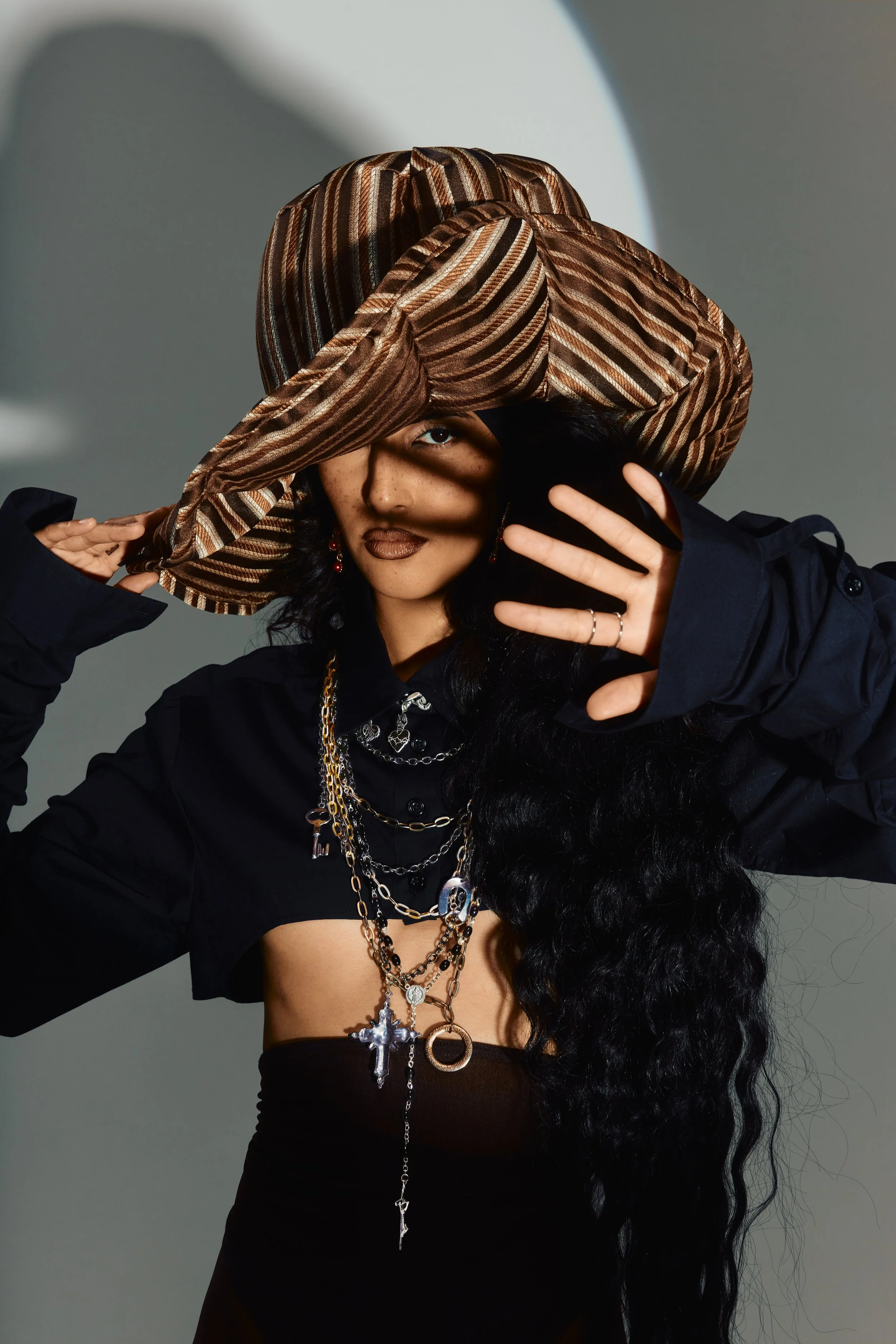 Styled by editorial stylist, Amanda Massi, a female model woman with long, curly black hair wearing multiple layered necklaces, a black top, and a large, wide-brimmed striped hat, posing with her hand partly covering her face, against a plain backgro