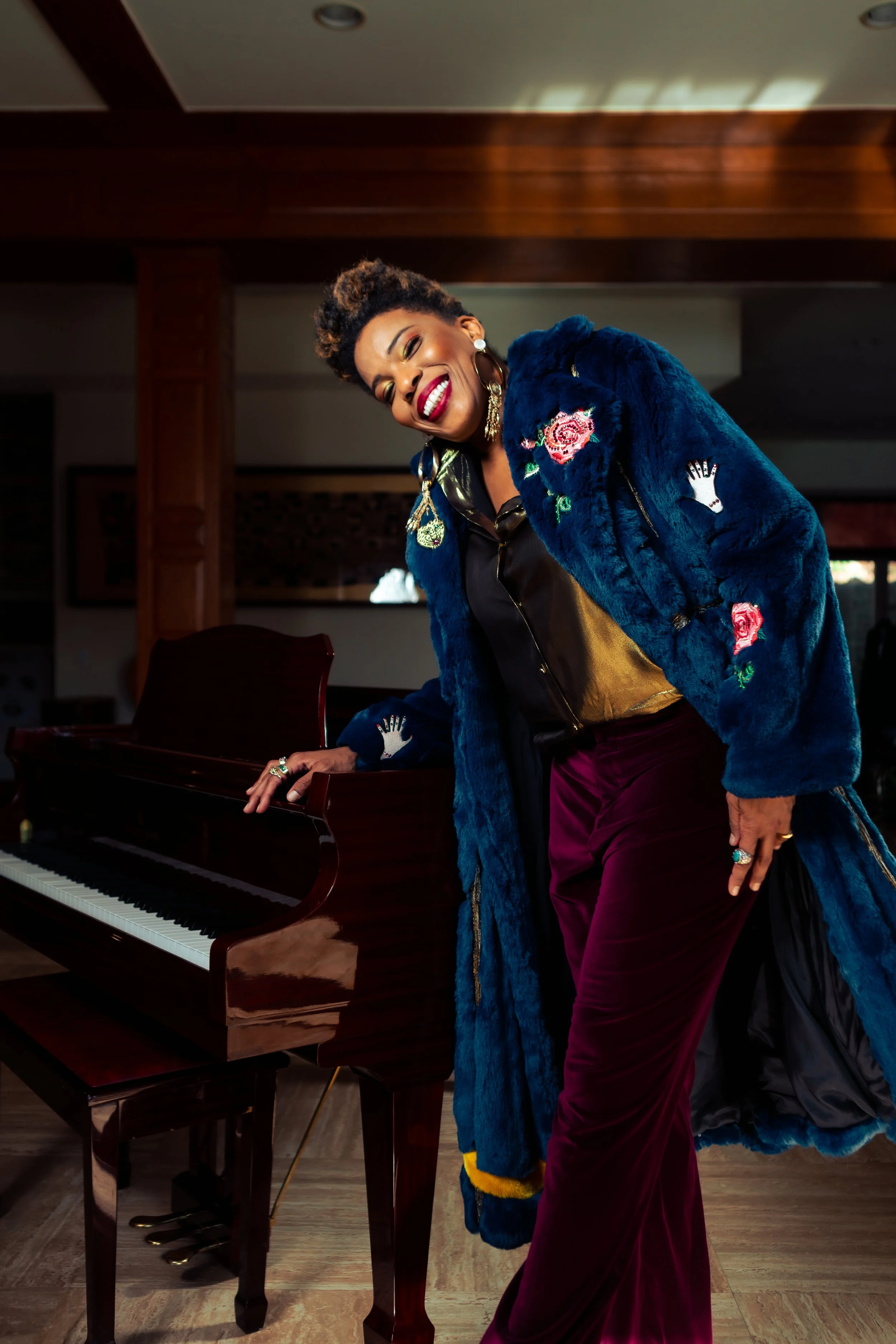 Styled by celebrity stylist, Amanda Massi, music icon Macy Gray wears a blue fur coat decorated with embroidered flowers and hand patches, standing next to a piano, indoors with wooden ceiling and floor for a fashion editorial in So FN Dope Magazine.