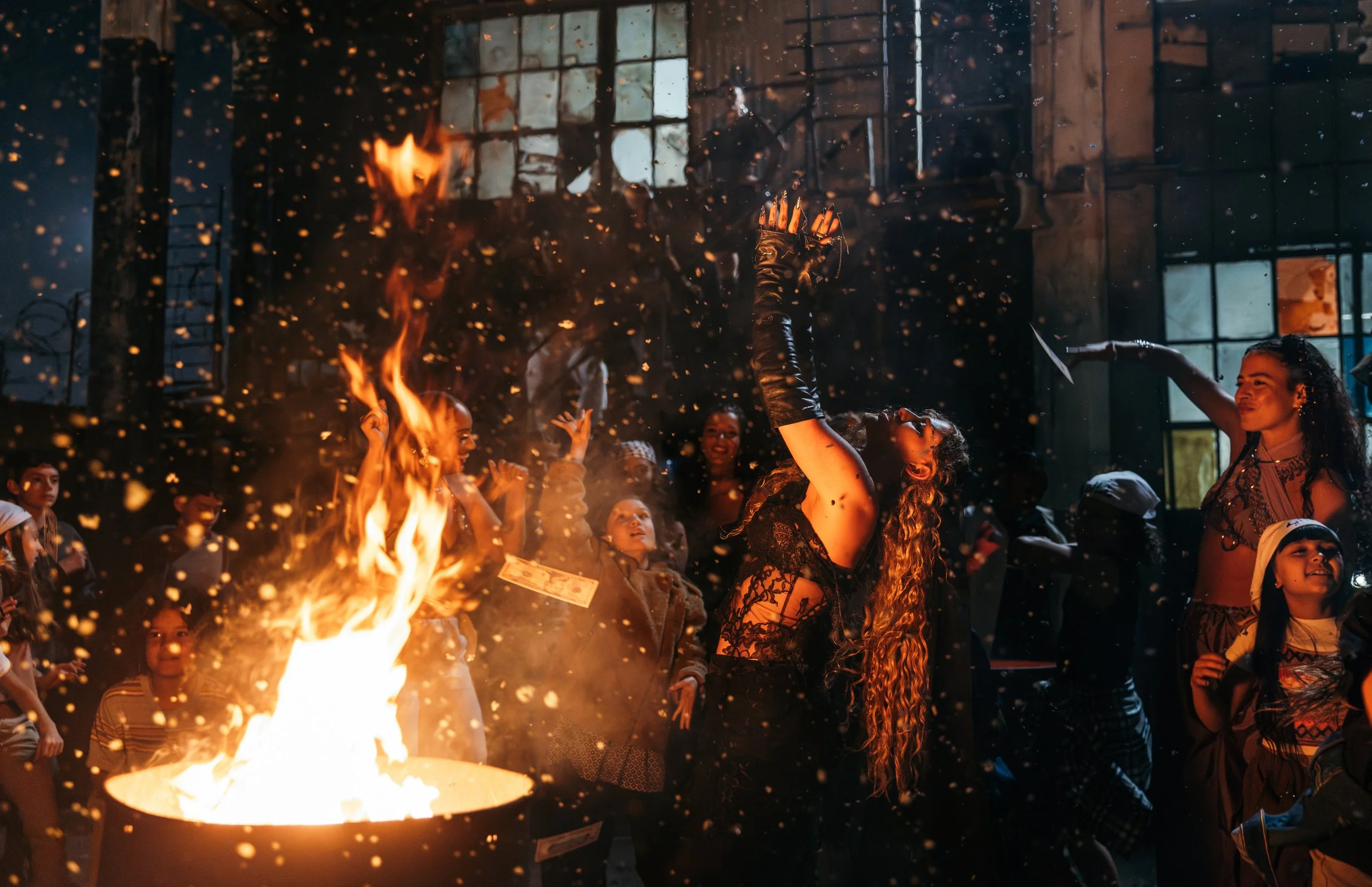 People gathered around a fire at night, with a woman holding a lit torch or candle, inside a building with large windows.