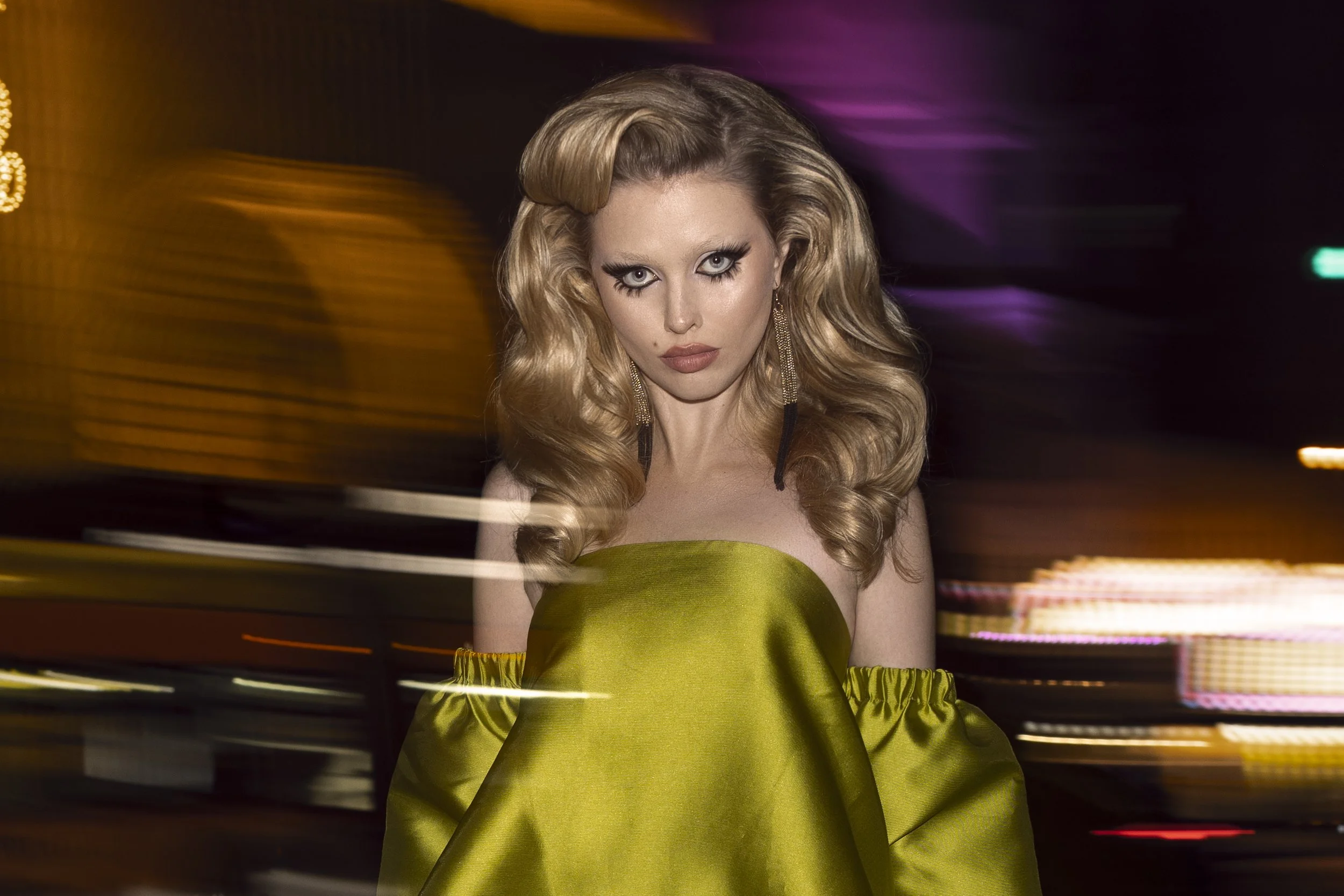Styled by editorial stylist, Amanda Massi, a female model with long, wavy blonde hair wearing a shiny, strapless green dress and long, dangling earrings, with a dramatic makeup look, in a setting with colorful blurred lights in the background for a f