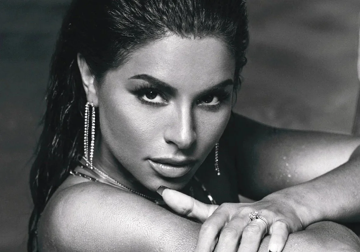 Black and white photo of a woman with wet hair and wet skin, looking intensely at the camera. She is wearing jewelry including earrings, a ring, and a necklace, and is resting her arm on a surface. The image includes the text "MAXIM" at the bottom left corner.