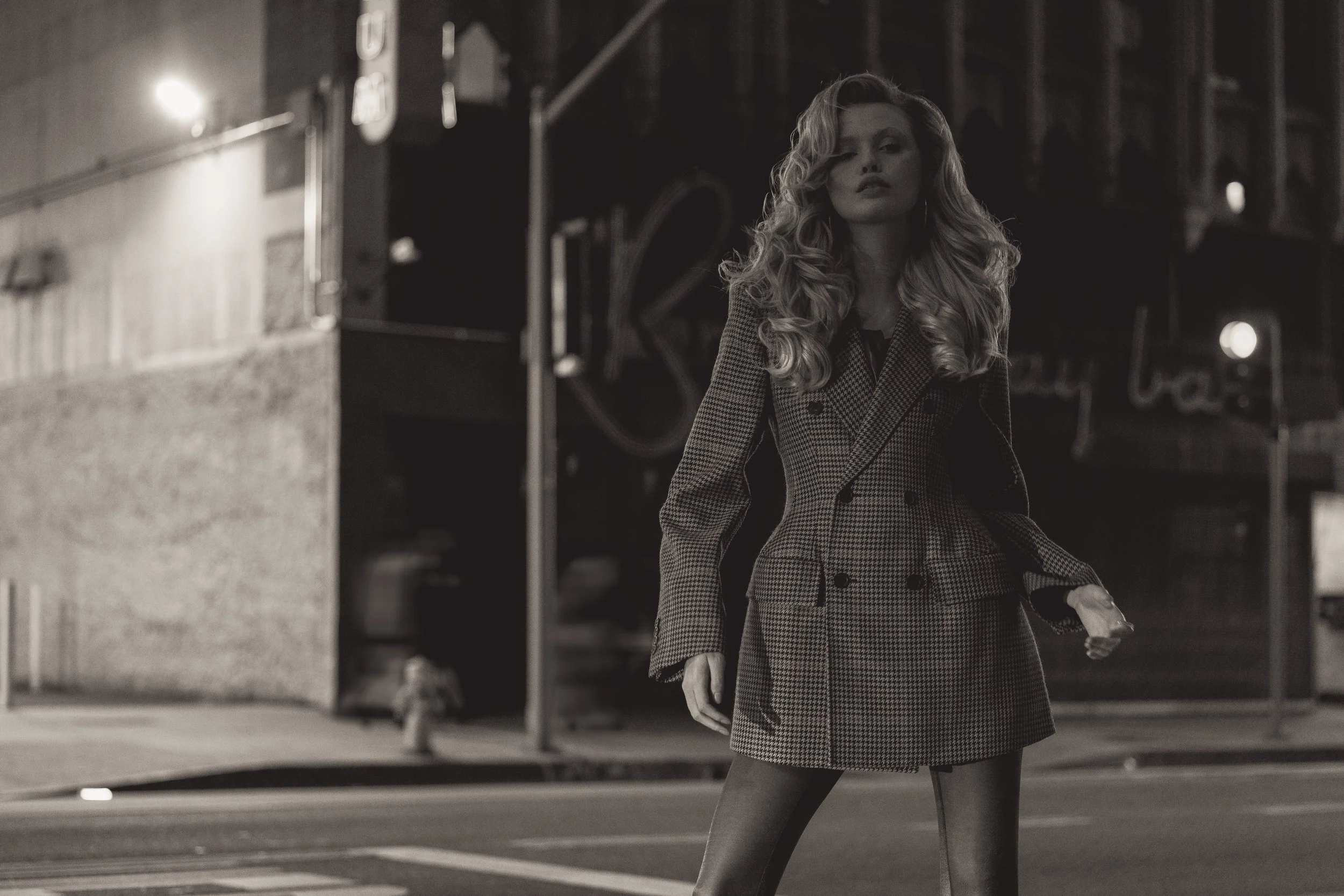 A woman with long, curly hair, wearing a checkered coat, standing on a city street at night, holding a purse and looking into the camera.