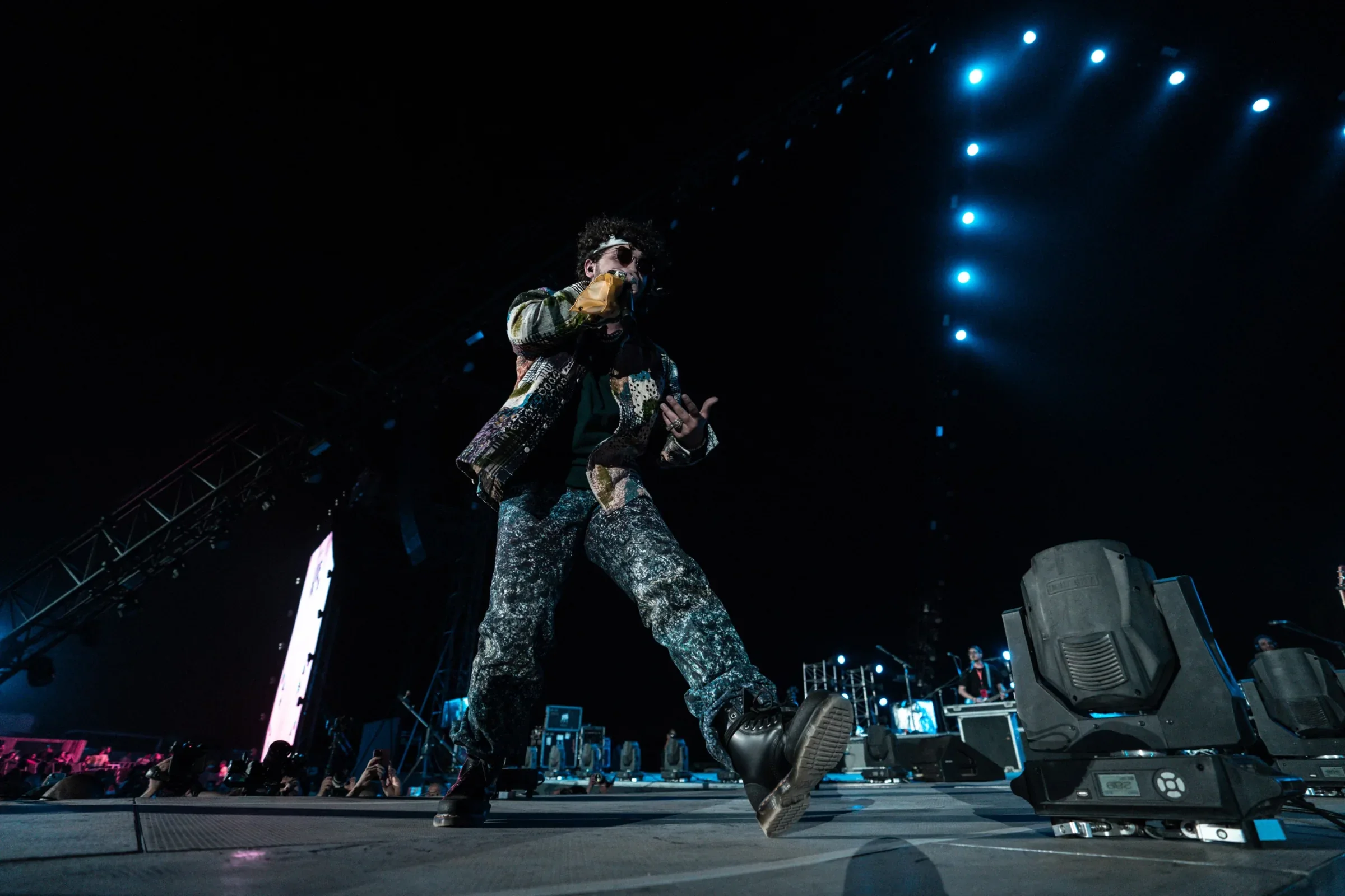 Styled by celebrity stylist, Amanda Massi, music artist Issam Alnajjar performs as the opening act for the Maroon 5 World Tour wearing a colorful outfit and sunglasses dancing on stage at a concert with blue stage lights overhead.