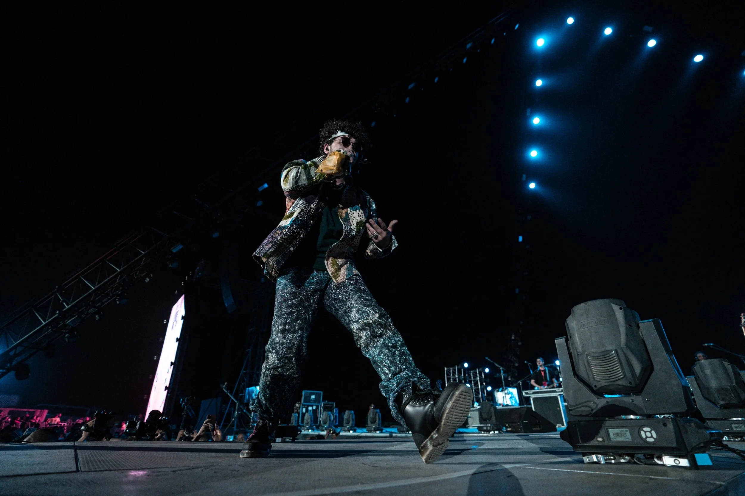 Styled by celebrity stylist, Amanda Massi, music artist Issam Alnajjar performs as the opening act for the Maroon 5 World Tour wearing a colorful outfit and sunglasses dancing on stage at a concert with blue stage lights overhead.