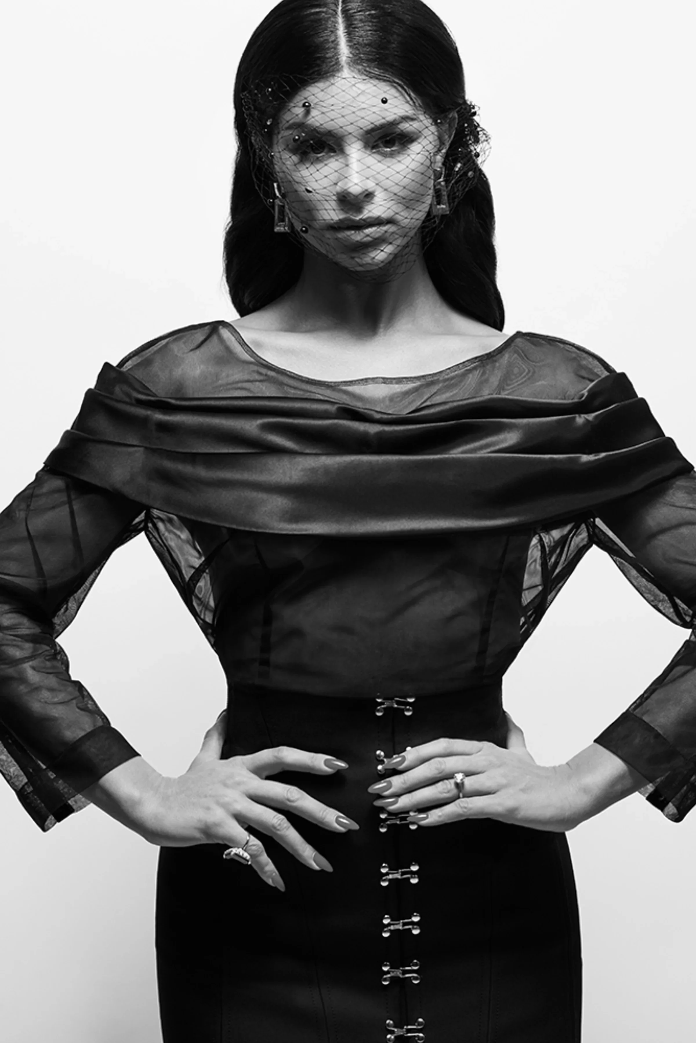 Styled by celebrity stylist, Amanda Massi, Rima Fakih Slaiby wearing a sheer top with layered fabric around the shoulders, a fitted high-waisted skirt with decorative buttons down the front, and a netted veil with small embellishments over her face. 