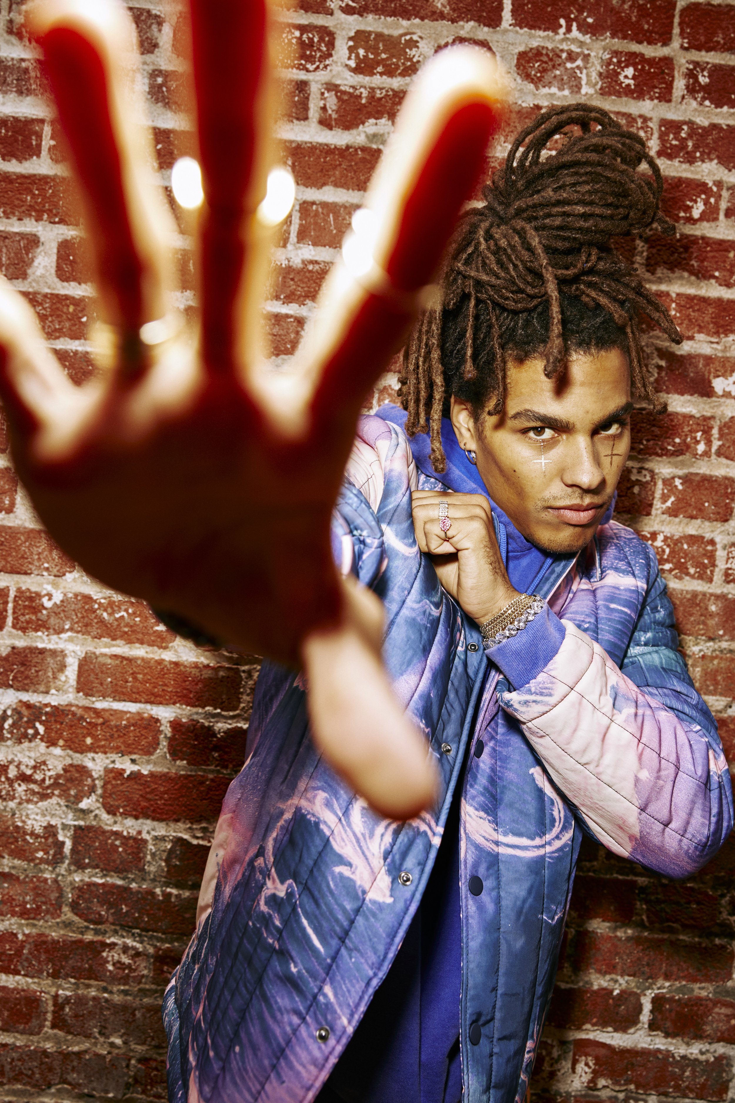 Styled by editorial stylist, Amanda Massi, a male model with dreadlocks and facial make-up wearing a colorful jacket, reaching out with their hand towards the camera, against a brick wall background for an editorial photoshoot.