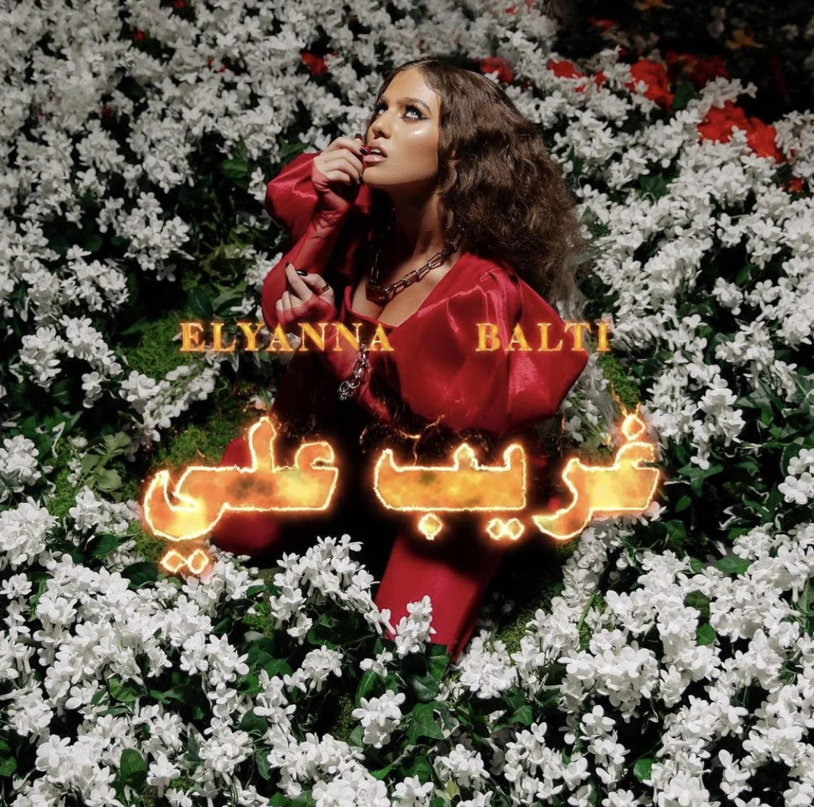 Styled by celebrity stylist and costume designer, Amanda Massi, music artist Elyanna wearing a custom red satin outfit, is sitting among white flowers with some red flowers in the background. There is glowing text in Arabic script, with the names 'El