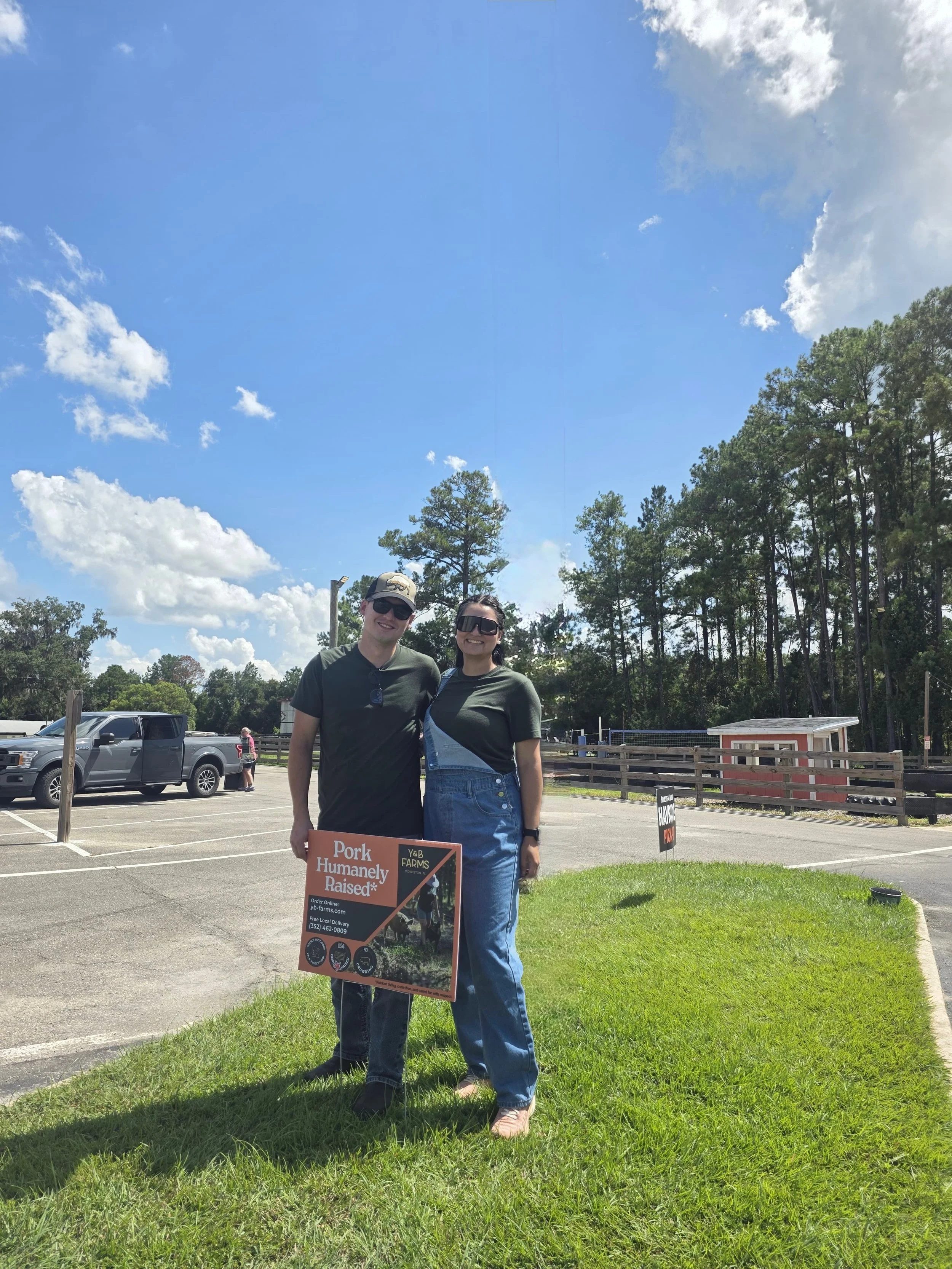 Yola and Bruno stand outdoors holding a Y&B Farms promotional sign on a sunny day, representing their pasture-raised pork farm at a local market.