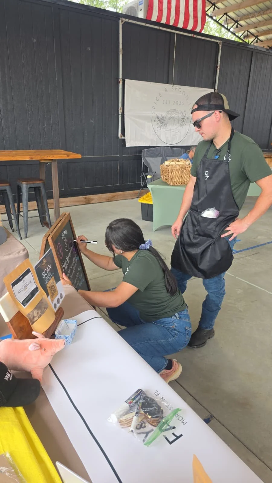 Yola prepares a market display by writing on a chalkboard sign at the Y&B Farms booth before customers arrive.