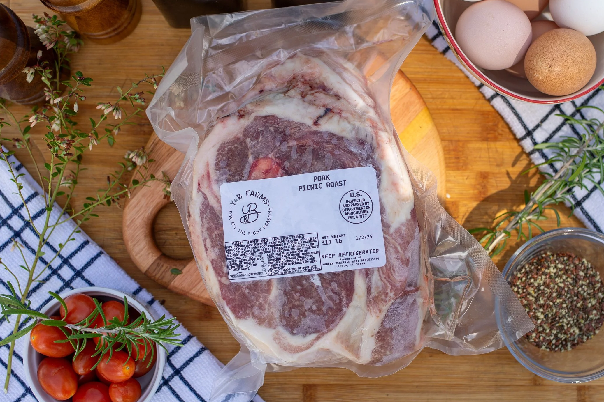 A vacuum-sealed package of pork picnic roast on a wooden cutting board, surrounded by eggs in a bowl, sprigs of rosemary, and small tomatoes, with a checkered cloth in the background.