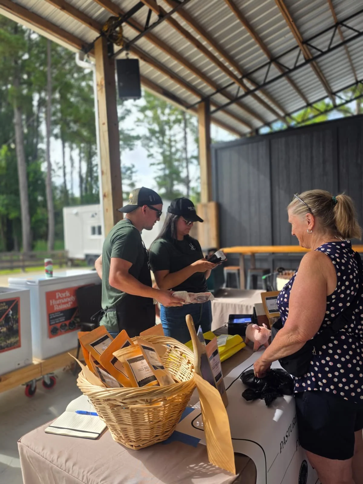 A customer speaks with two Y&B Farms vendors at a market booth. Packaged pet treats are displayed in baskets on the table, and freezers containing farm products are positioned behind them inside a covered outdoor market space.