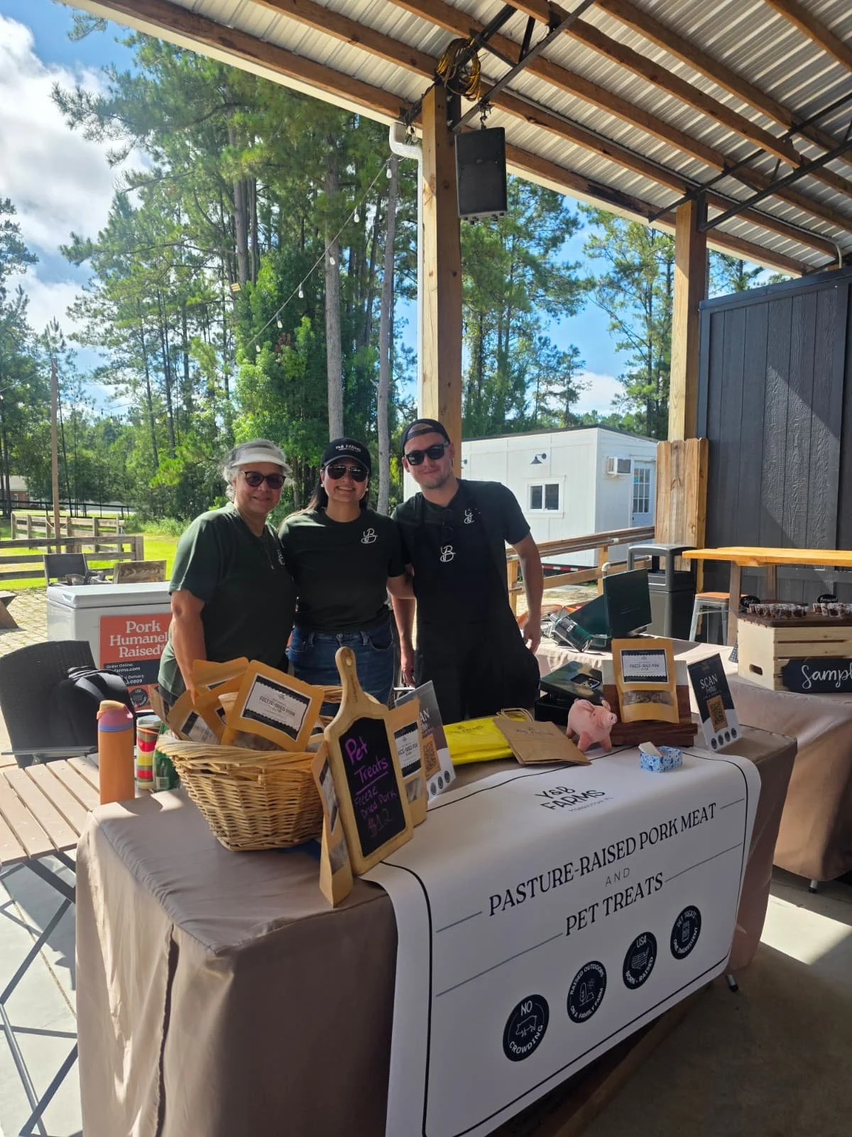 Y&B Farms vendors stand behind a market table displaying freeze-dried pet treats in baskets while assisting a customer. The booth is set up inside a covered farmers market pavilion with pork products stored in freezers nearby.