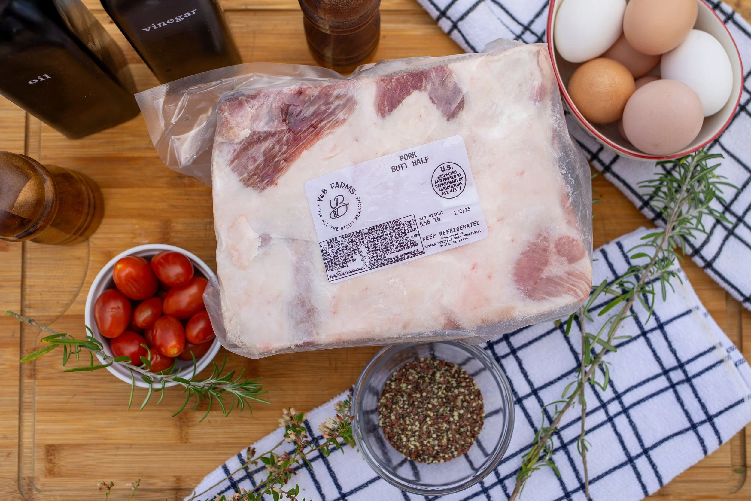 Pasture-raised pork butt from Y&B Farms, USDA-inspected and vacuum-sealed, displayed with fresh tomatoes and herbs.