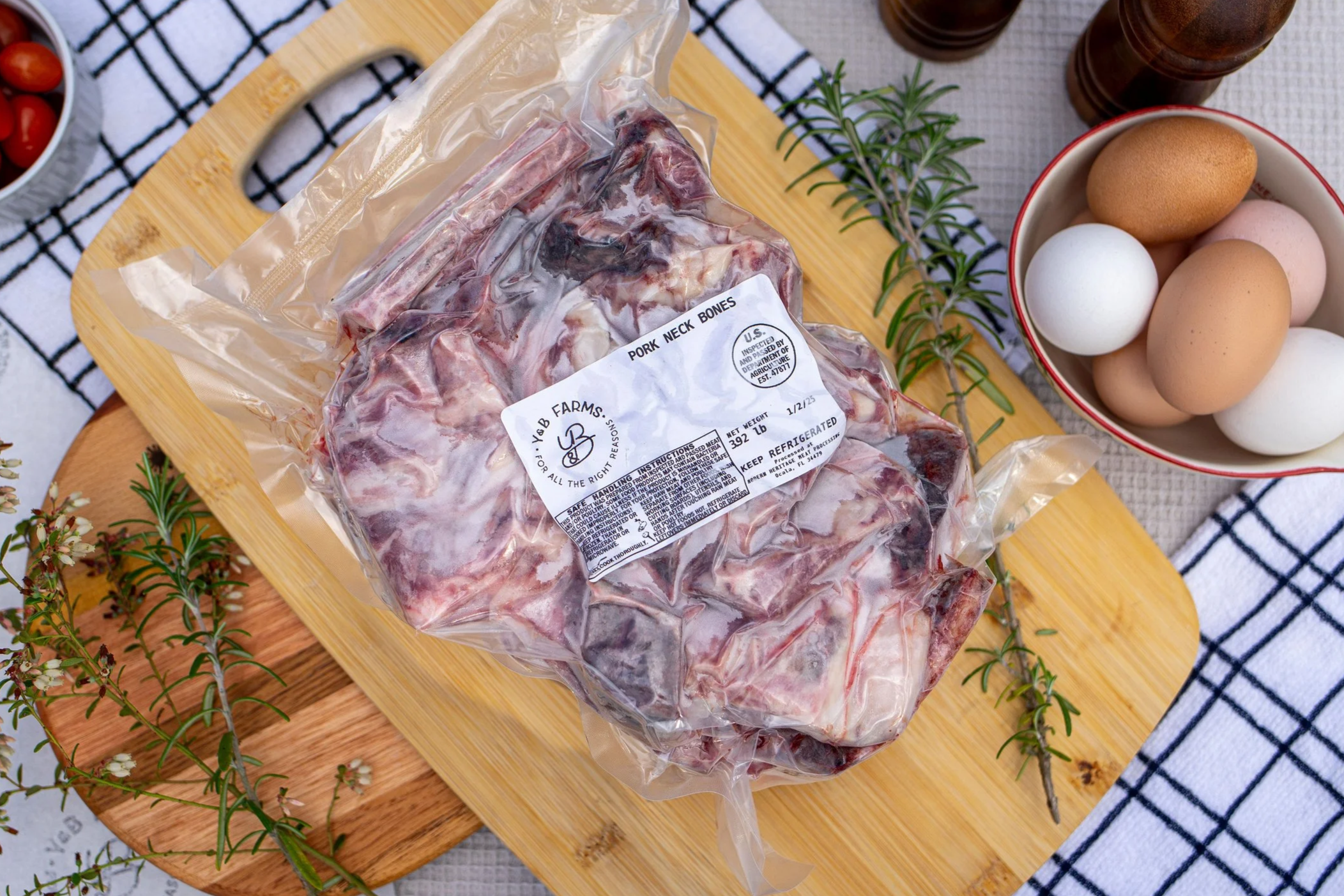 A vacuum-sealed package of pork neck bones on a wooden cutting board, surrounded by eggs in a bowl, sprigs of rosemary, and small tomatoes, with a checkered cloth in the background.