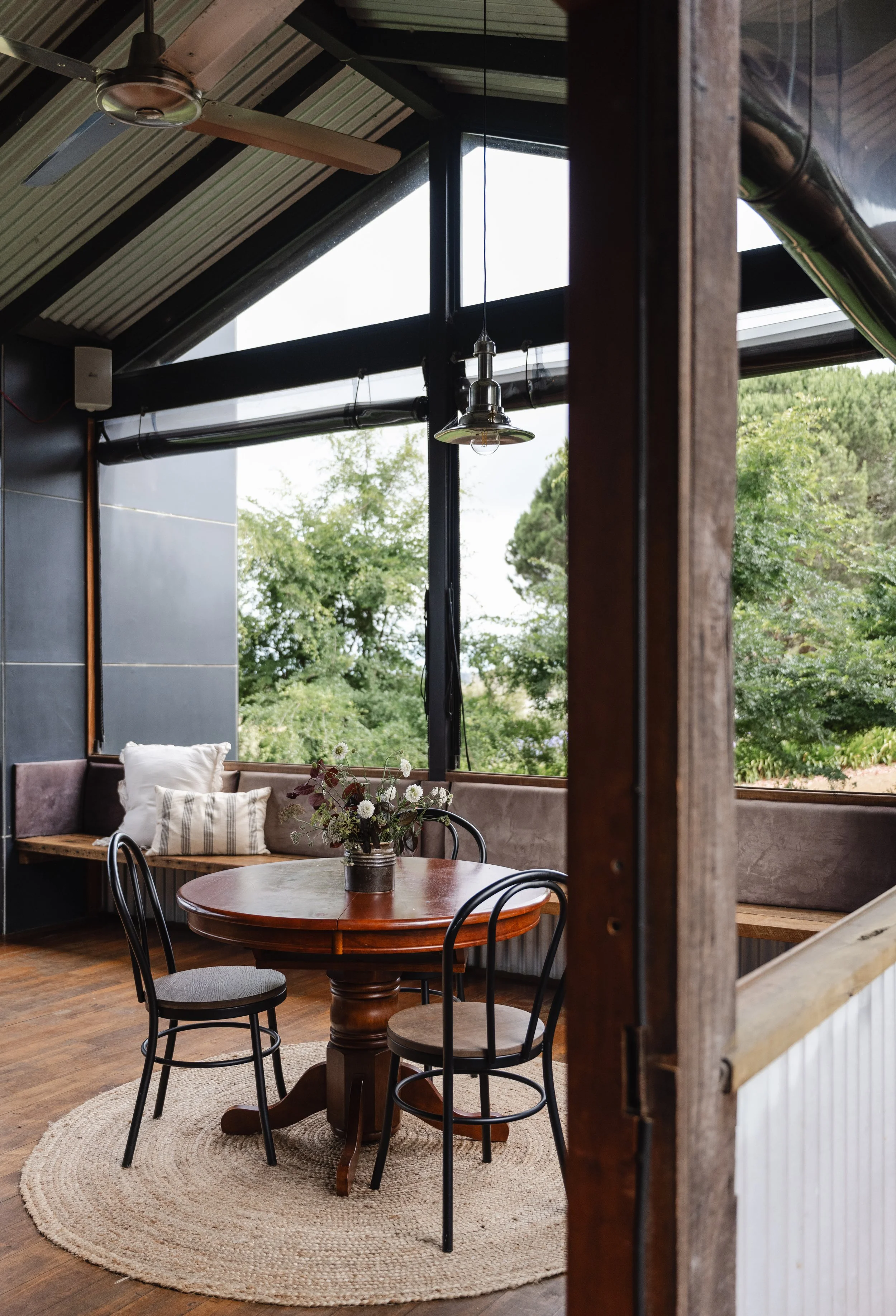 Interior of a cozy dining area with a round wooden table, black metal chairs, a flower arrangement, large windows showing greenery outside, and a woven rug on wooden flooring.