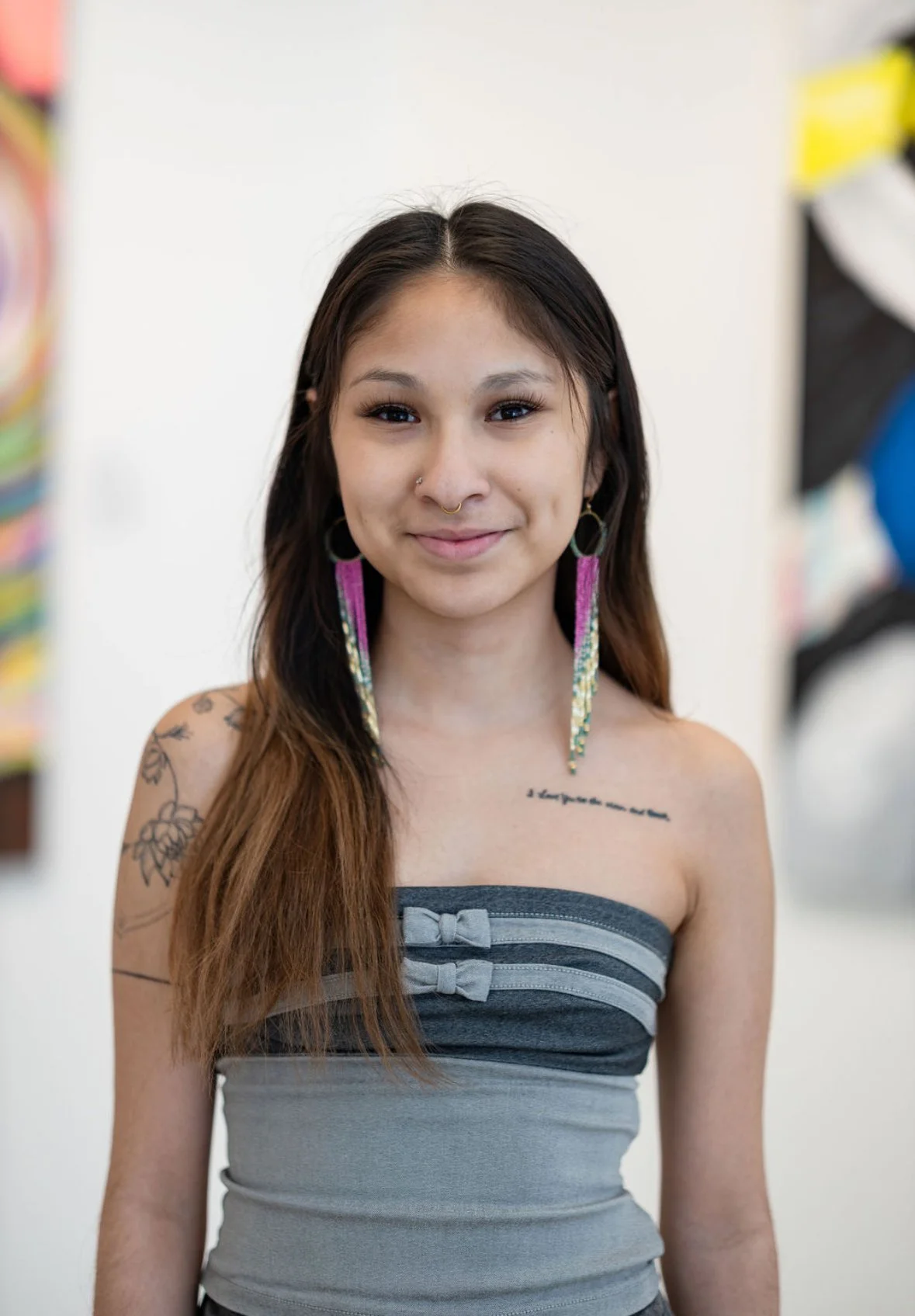 A young woman with long brown hair and a nose piercing is smiling at the camera. She is wearing large, colorful earrings and a sleeveless gray top with bow details.