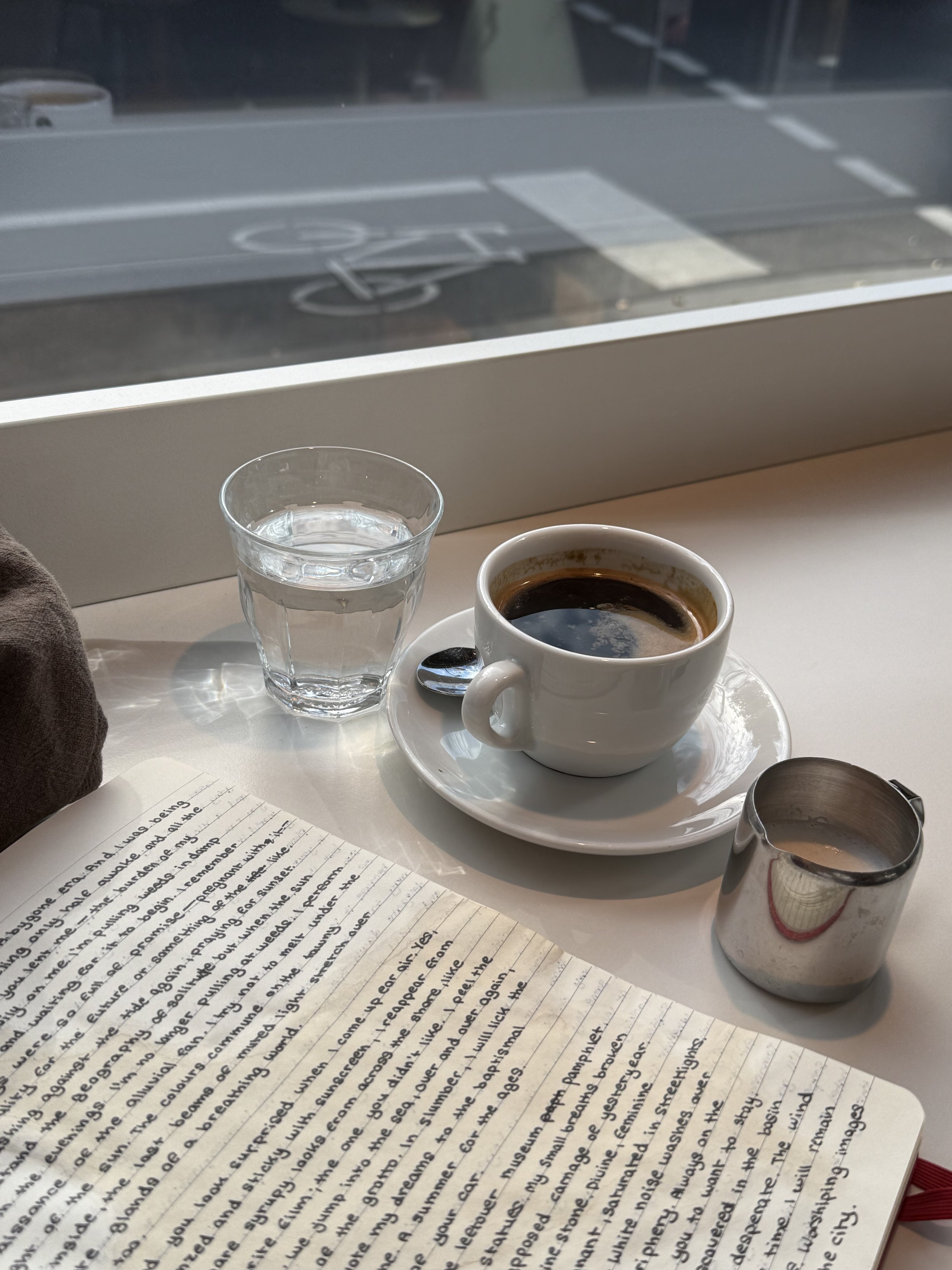 A cup of black coffee on a white saucer with a small spoon, a glass of water, a metallic creamer, a spoon on a white table near a window, and an open notebook with handwritten notes.