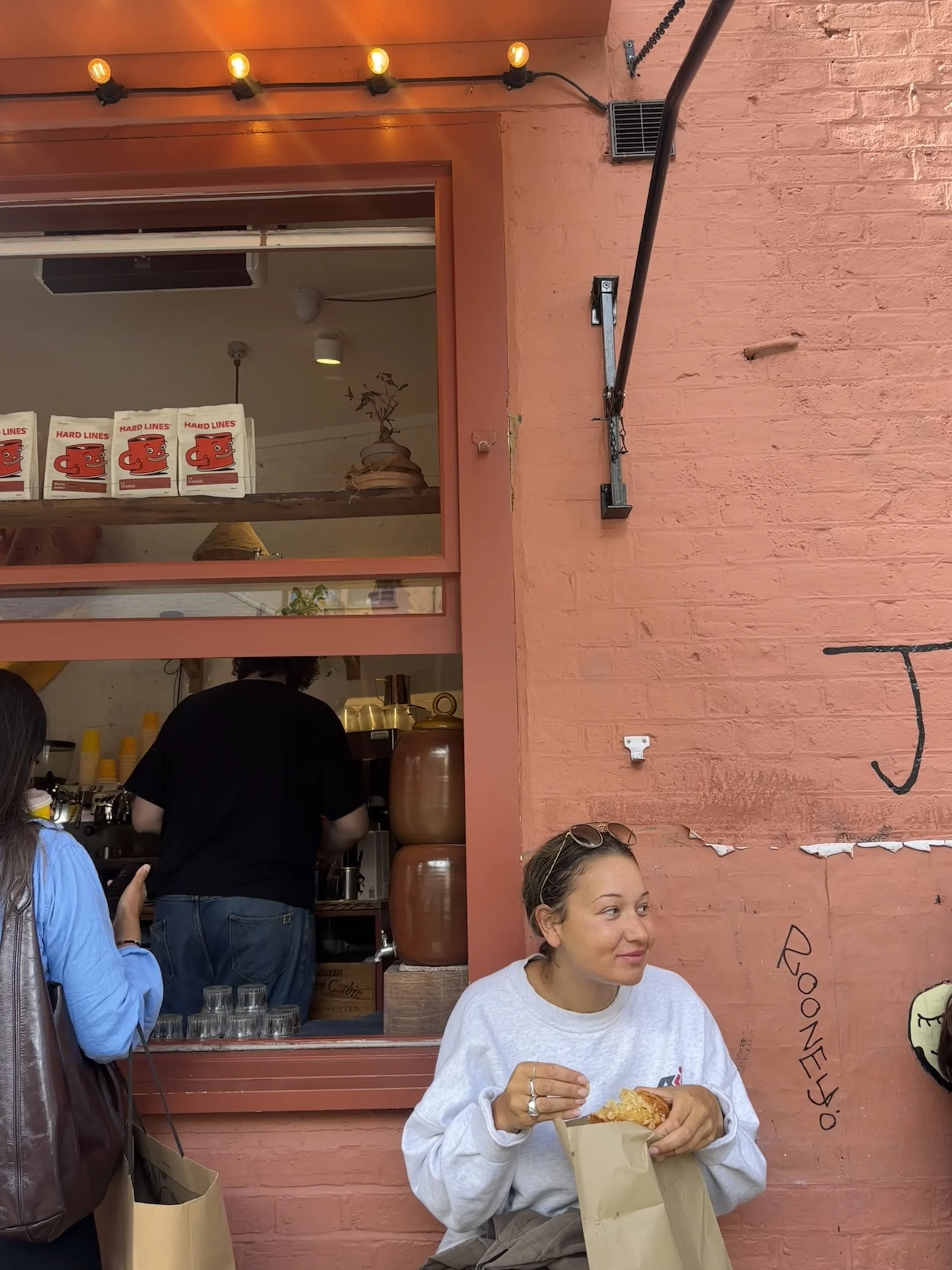 A woman sitting outside a restaurant, eating a slice of pizza. Behind her, a window shows the interior with a barista and kitchen equipment. The exterior wall is painted red and decorated with black graffiti.