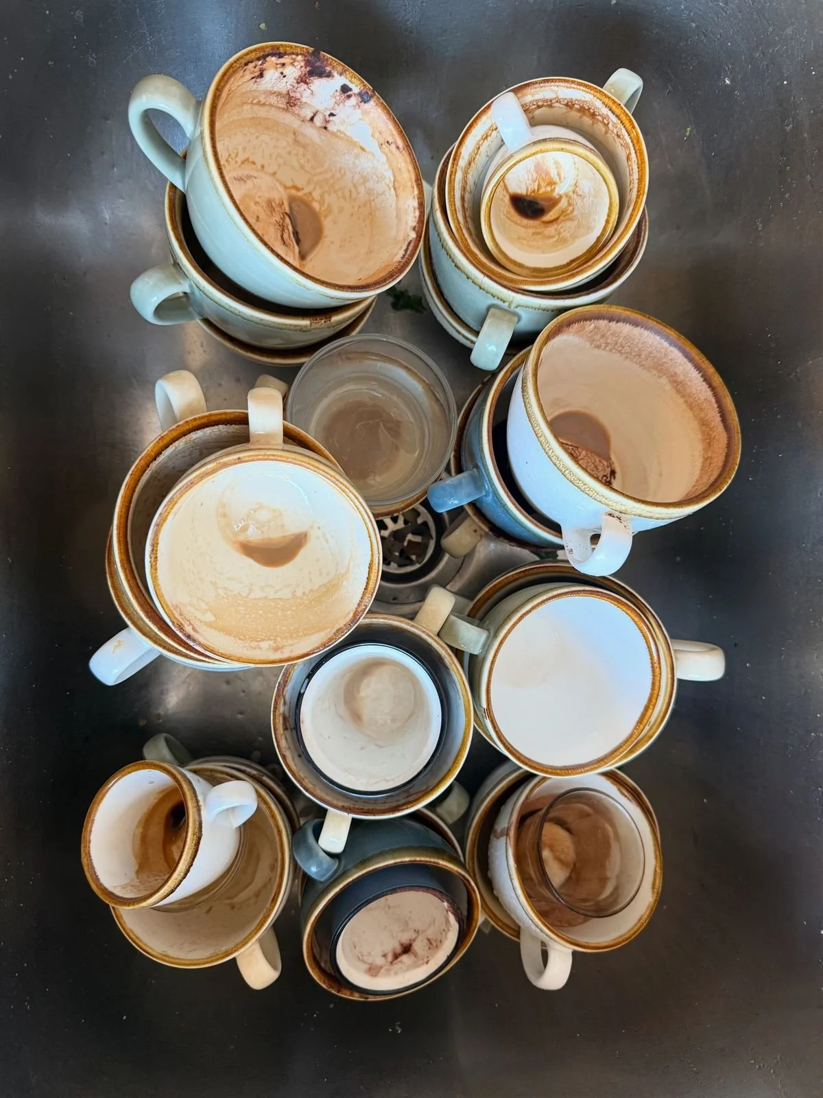 Multiple coffee cups stacked and placed together on a metallic surface, some filled with coffee remnants and some with liquid, with a small glass of water in the center.