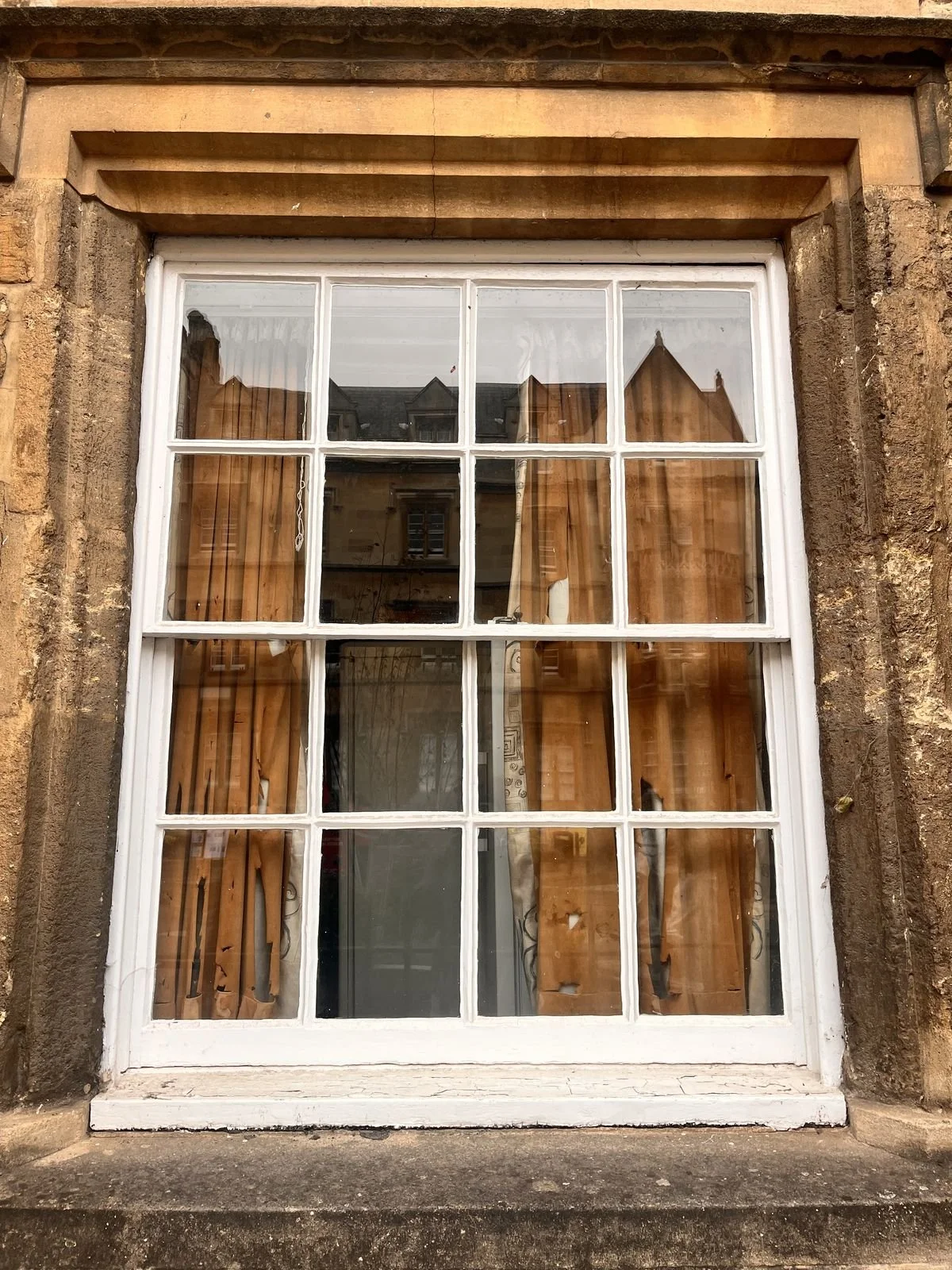A white-framed window with mullions, set in a weathered brick wall with tan keystone trim, showing curtains inside and reflections of neighboring buildings.