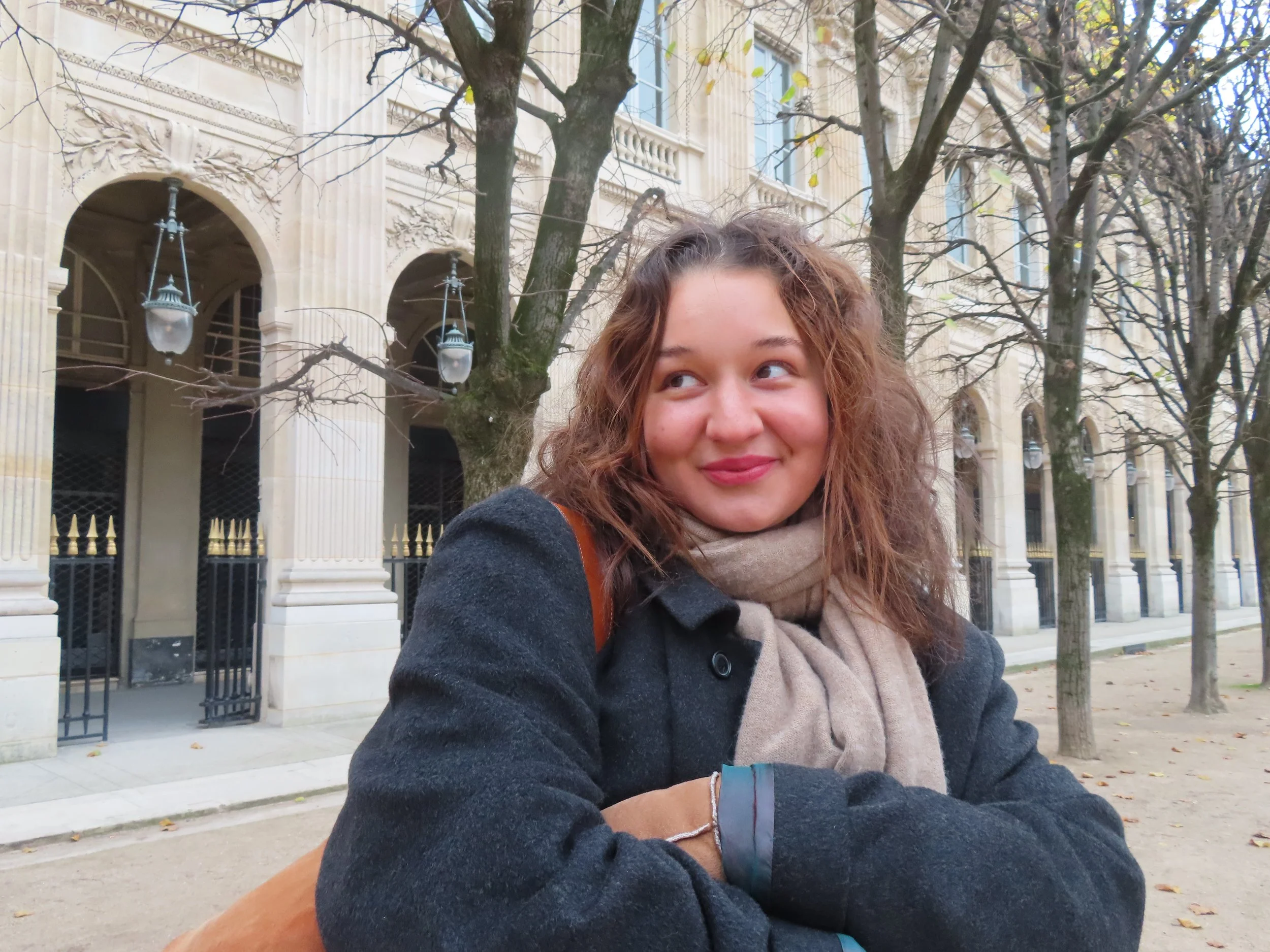 A young woman with curly hair wearing a dark coat and light scarf, sitting outdoors near leafless trees with an elegant building in the background.