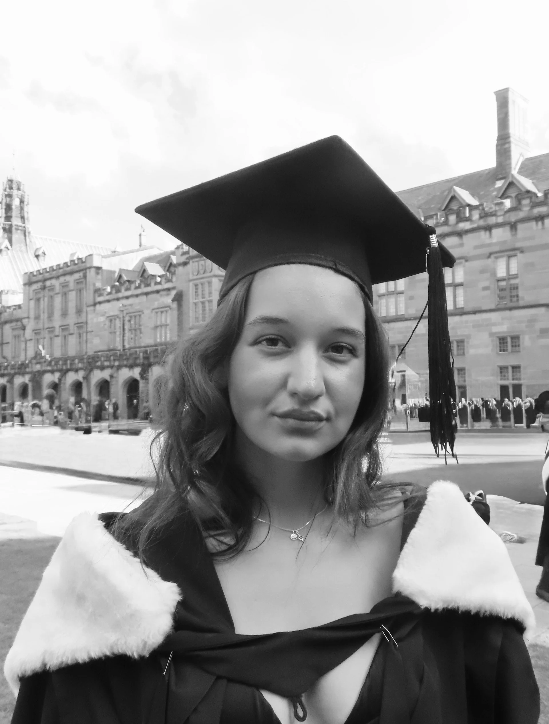 Young woman in graduation cap and gown outdoors in front of historic buildings.