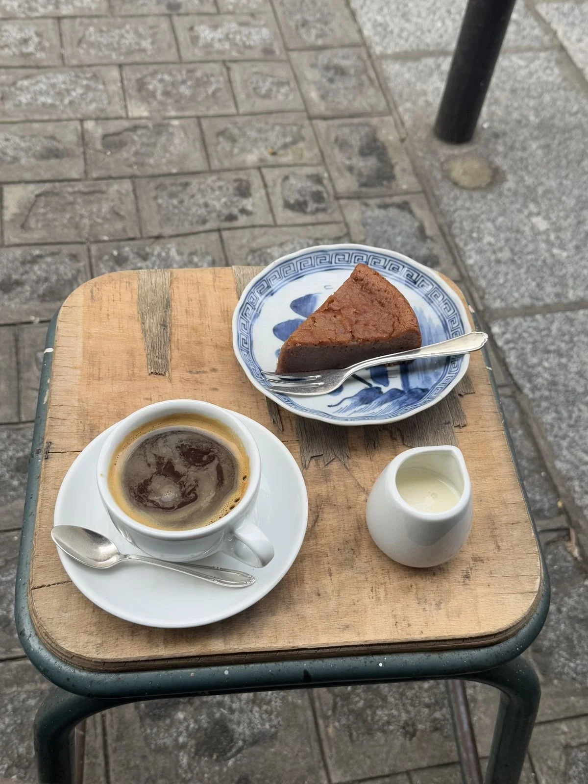 A cup of black coffee with a small spoon on a white saucer, a slice of chocolate cake on a blue and white ceramic plate with a fork, and a small white pitcher of cream on a wooden table outdoors.