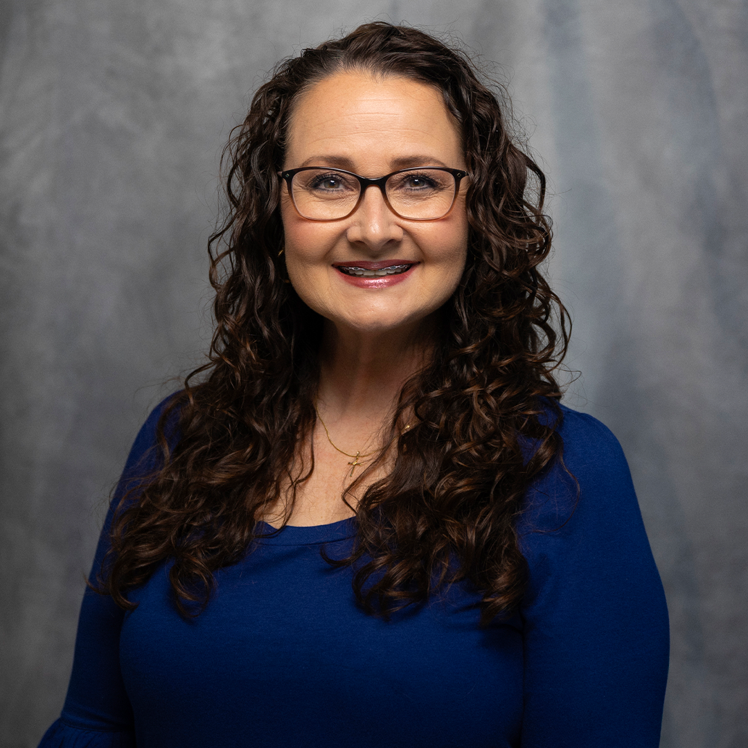 A woman with long, curly brown hair, smiling, wearing a teal ribbed sweater, against a gray background.