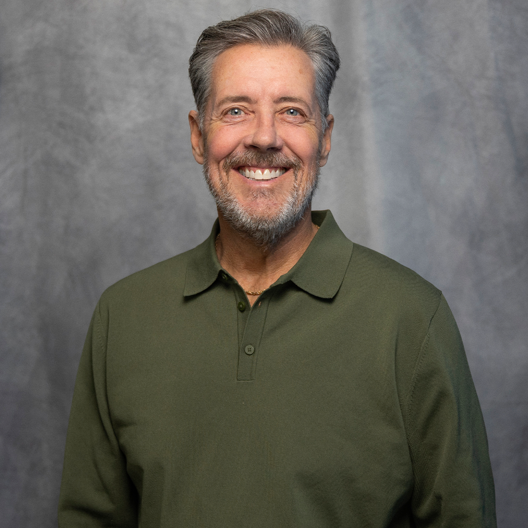 Smiling middle-aged man with gray hair and beard wearing a colorful, patterned plaid shirt against a gray background.