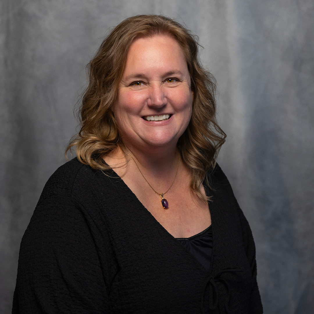 A woman with long wavy light brown hair, wearing a floral blouse with pink, green, and beige colors, and a gold necklace with a dark pendant, smiling at the camera against a gray gradient background.