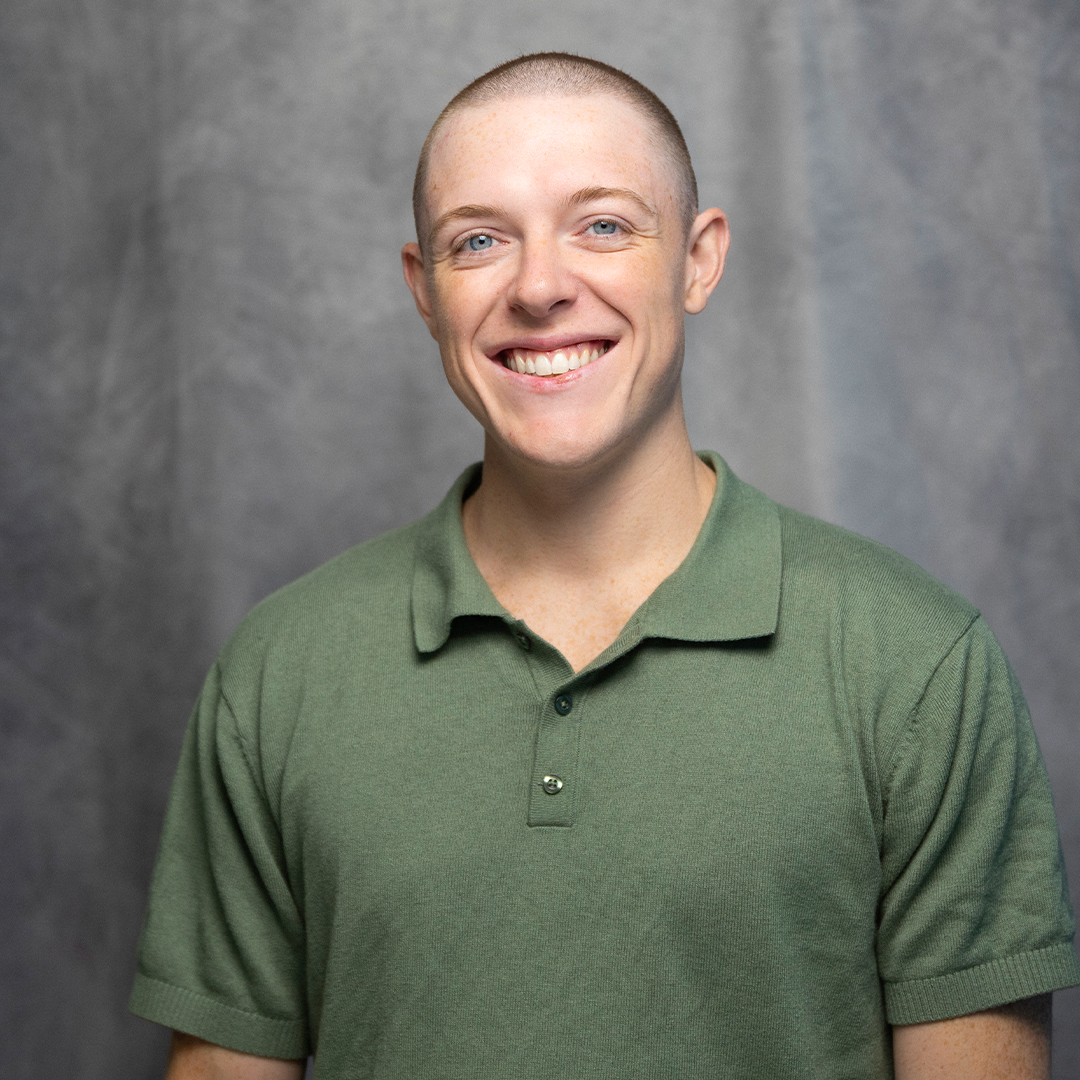A young man with short brown hair, blue eyes, and a light beard smiling, wearing a gray shirt, against a plain gray background.