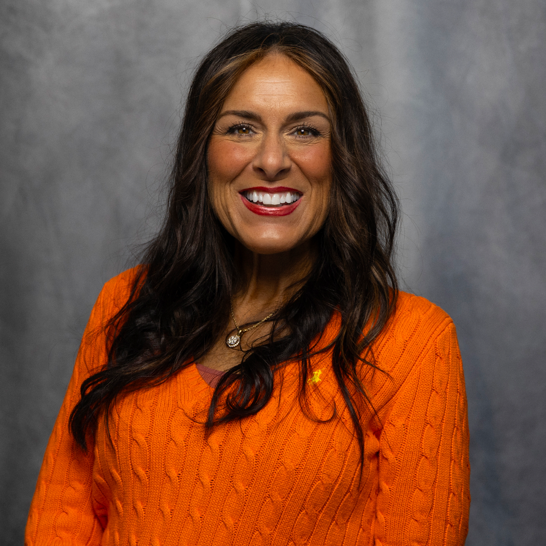 A woman with long black hair, wearing an orange cable-knit sweater and a gold necklace with a pendant, smiling against a neutral gray background.