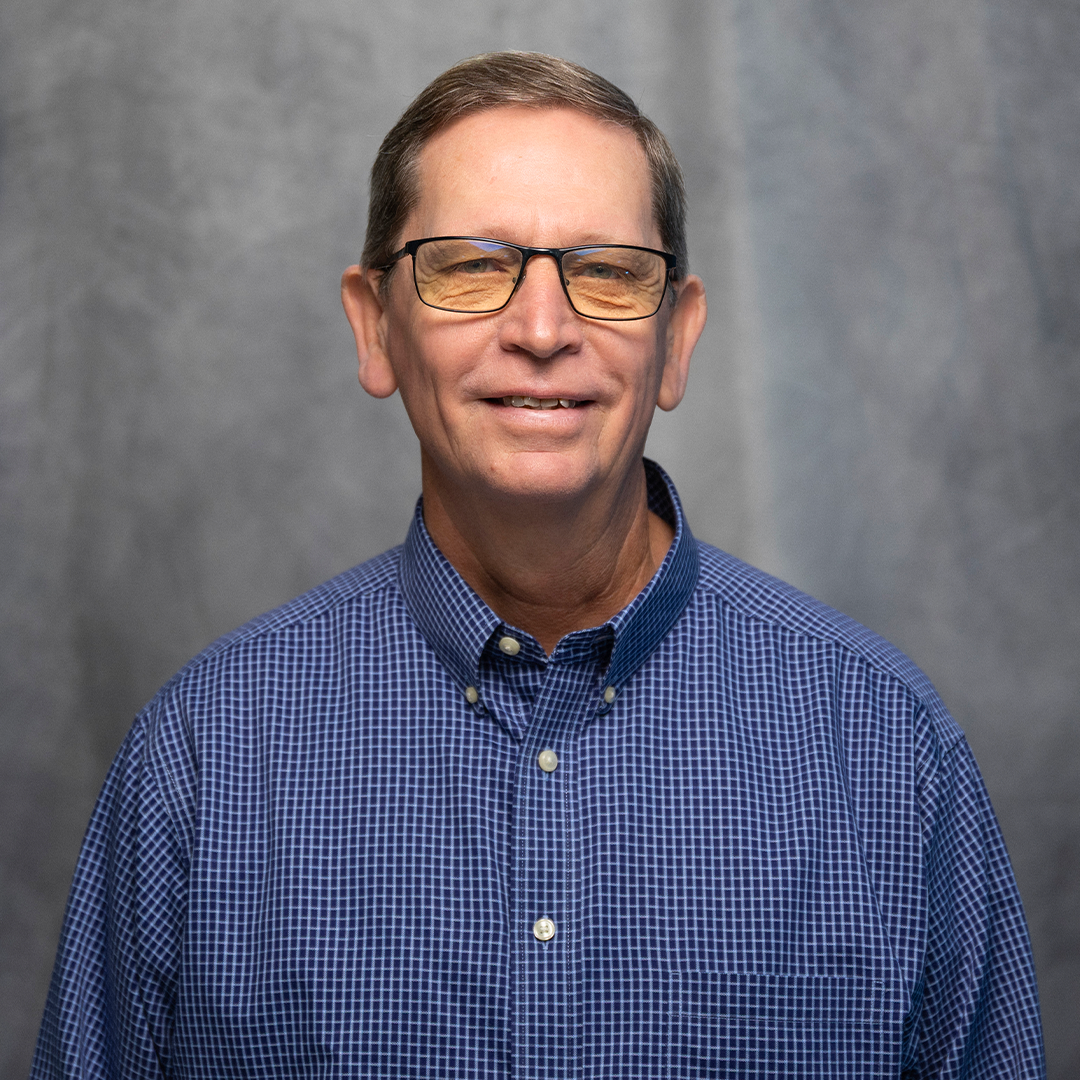 A smiling middle-aged man wearing glasses, a red checkered shirt, and a black vest against a plain background.