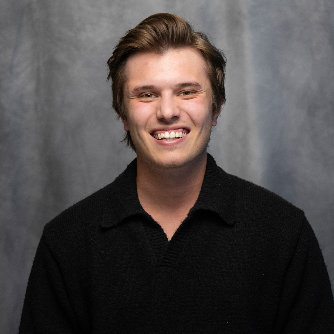 Young man with brown hair smiling, wearing a brown t-shirt and black jacket, against a gray background.