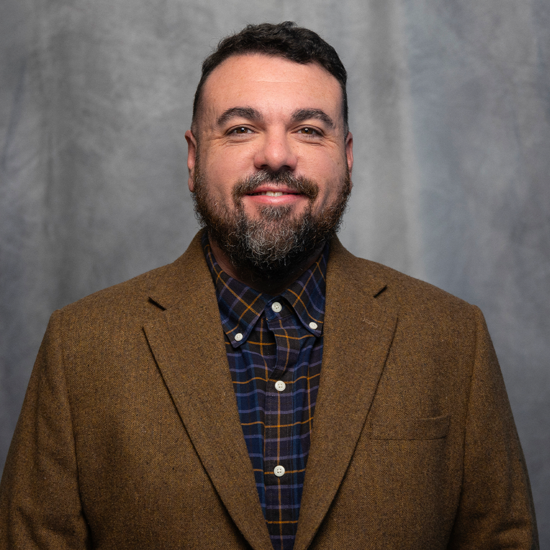 Headshot of a smiling man with dark, curly hair and a beard, wearing a light-colored button-up shirt over a black T-shirt, against a gray background.