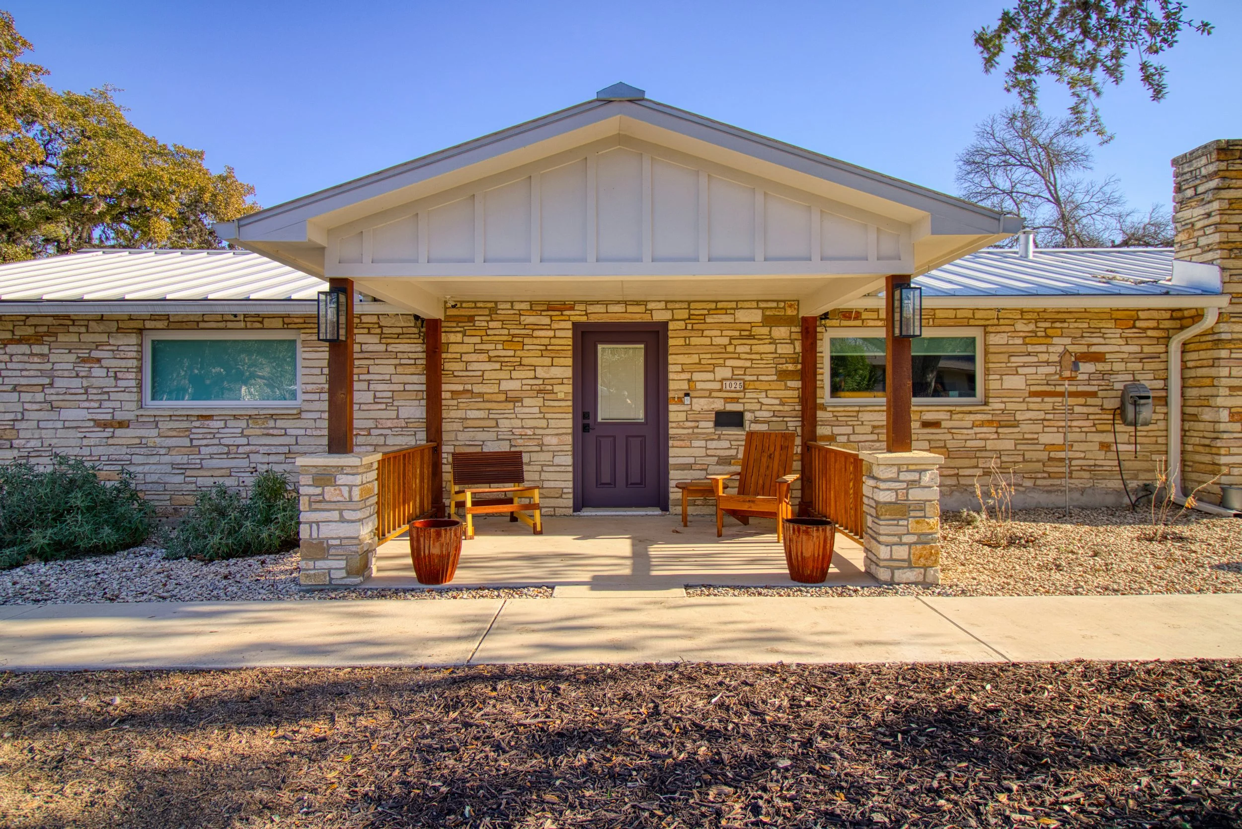 Front of a house with a small porch, two wooden chairs, and potted plants. The house has a stone exterior, a purple door, and a white metal roof.