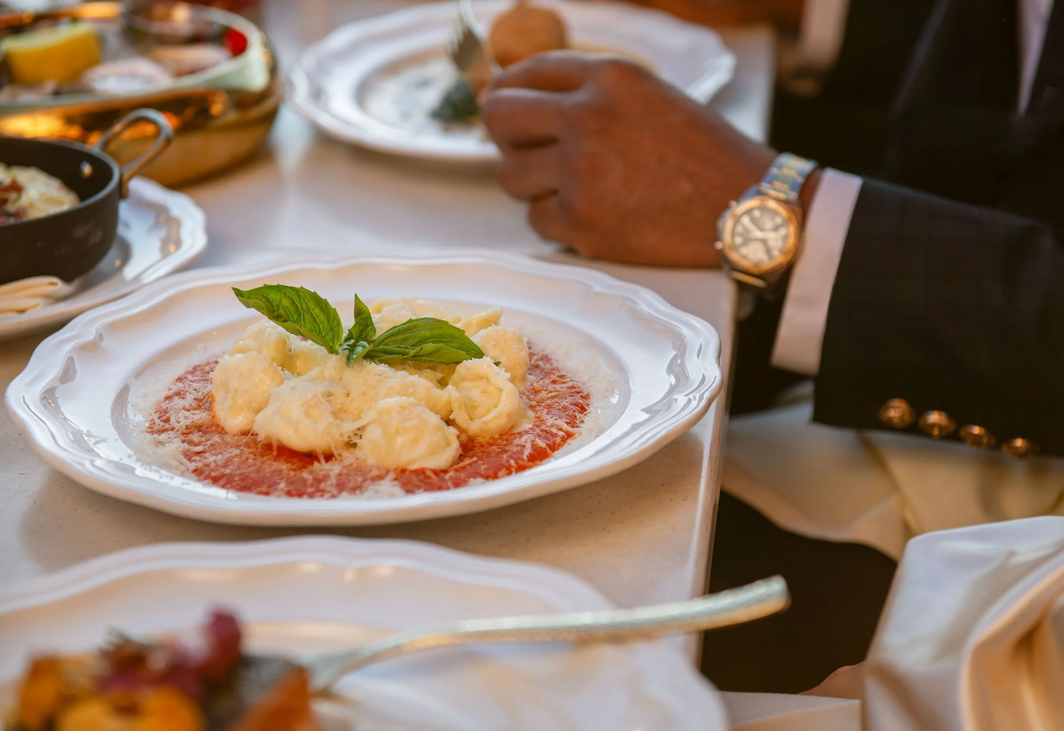 A white ornate plate with a serving of spaghetti topped with cheese and garnished with basil, set on a dining table  at Cheri Rooftop with other dishes and a person's hand reaching for food.