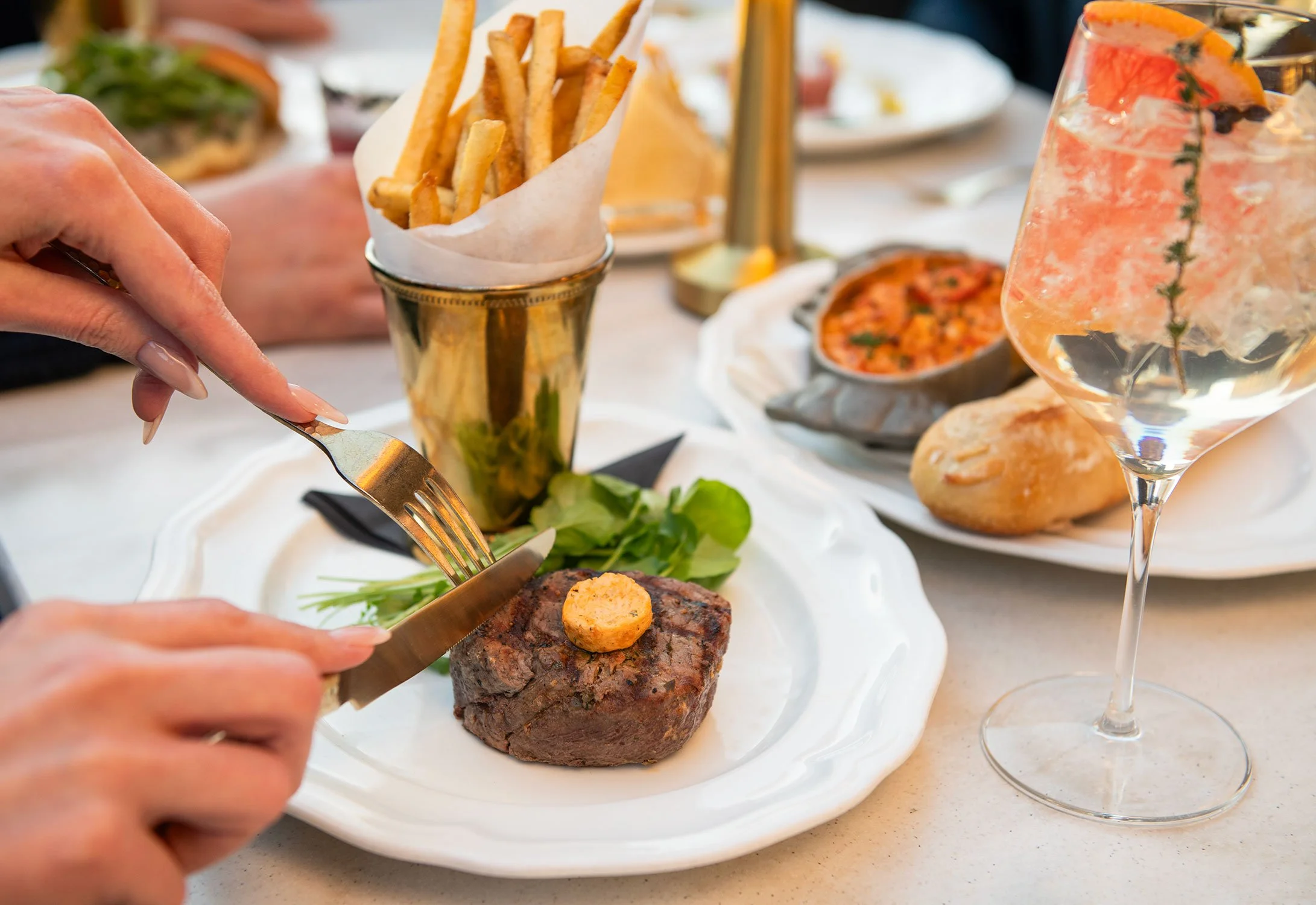A plate with a cooked steak topped with a pat of butter, set on a table with assorted dishes including french fries, salad, pizza, and a glass of water with garnishes  at Cheri Rooftop.